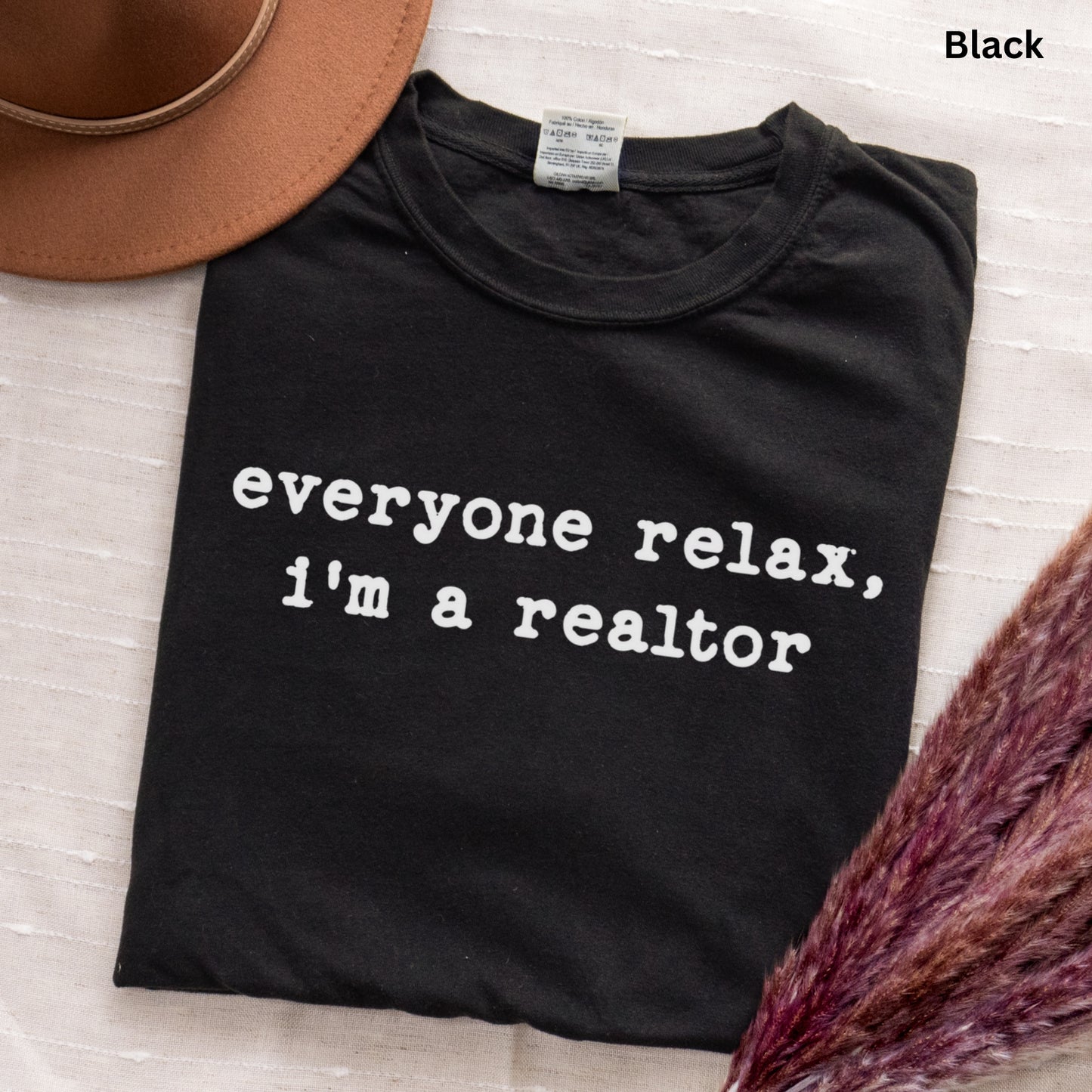 A black t-shirt with white text that says "everyone relax, i'm a realtor" is displayed on a white surface, accompanied by a brown hat and some purple flowers.