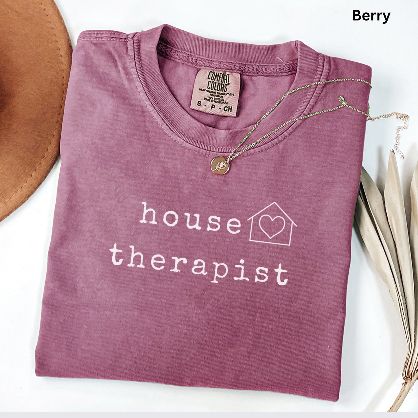 A pink t-shirt with the text "house therapist" printed on it, along with a heart symbol, is displayed on a white surface.