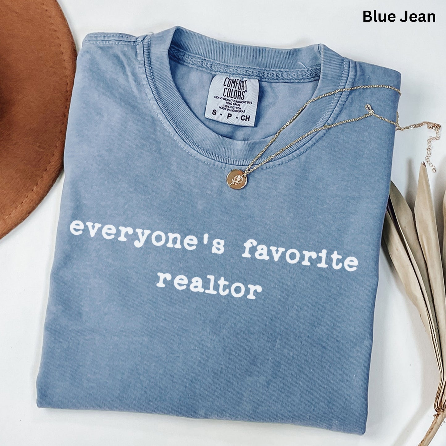 A blue t-shirt with the text "Everyone's favorite realtor" printed on it, along with a necklace and some leaves.
