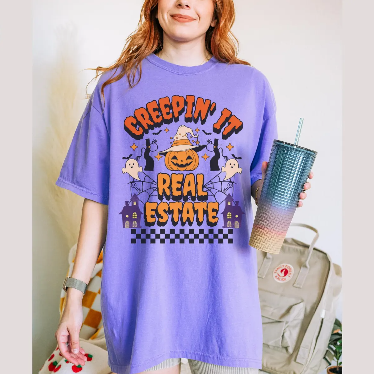 A woman wearing a purple t-shirt with a Halloween-themed graphic and text, holding a drink in a cup.