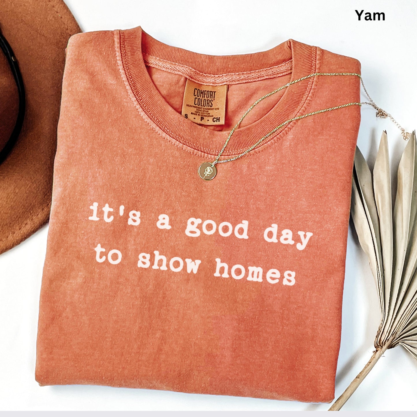 a red t-shirt with the text "It's a good day to show homes" printed on it, along with a gold necklace and a brown hat.