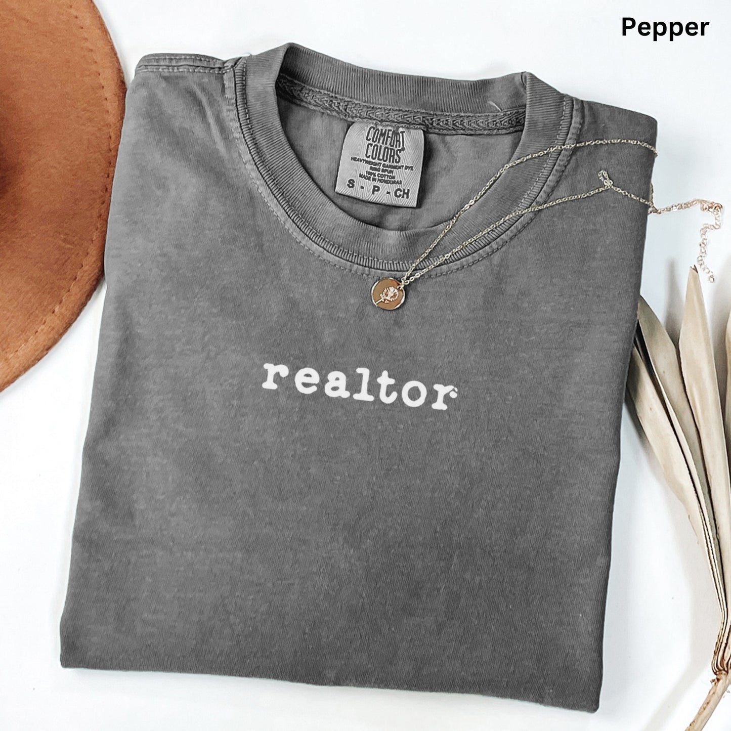 A gray t-shirt with the word "realtor" printed on it, along with a necklace and a hat, all laid out on a white surface.