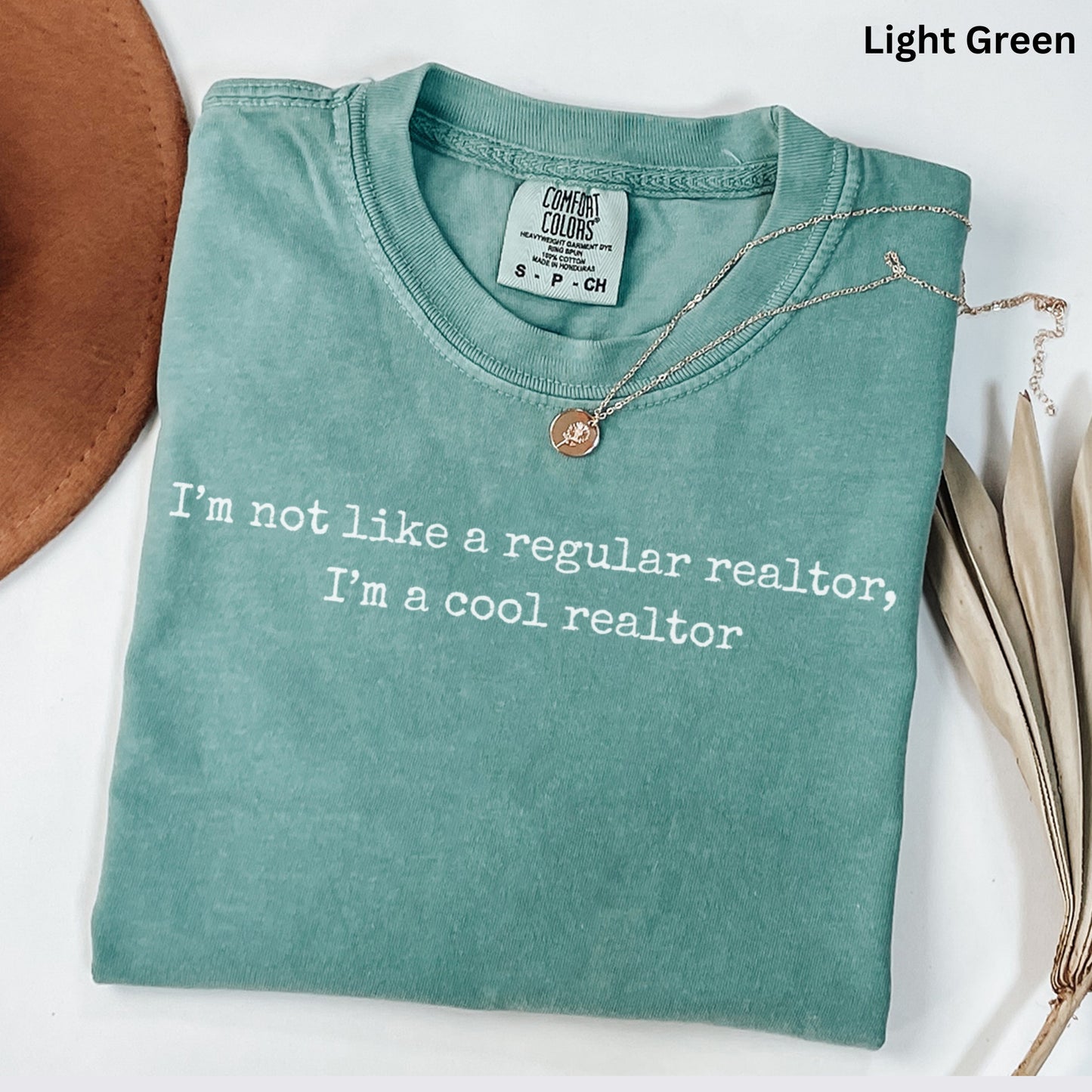A light green t-shirt with a humorous text printed on it, along with a necklace and some dried leaves, all laid out on a white surface.