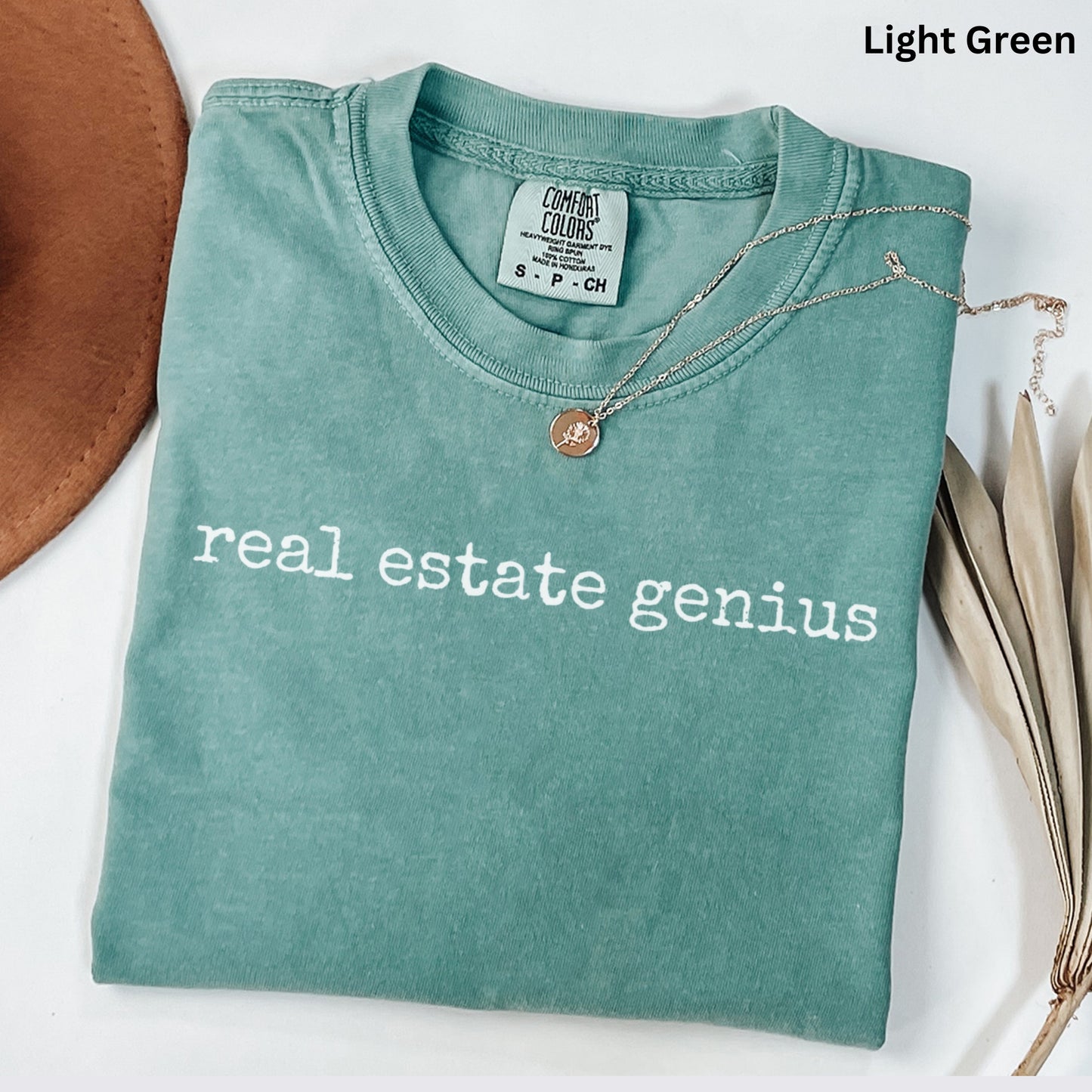 A light green t-shirt with the text "real estate genius" printed on it, along with a necklace and a brown hat.
