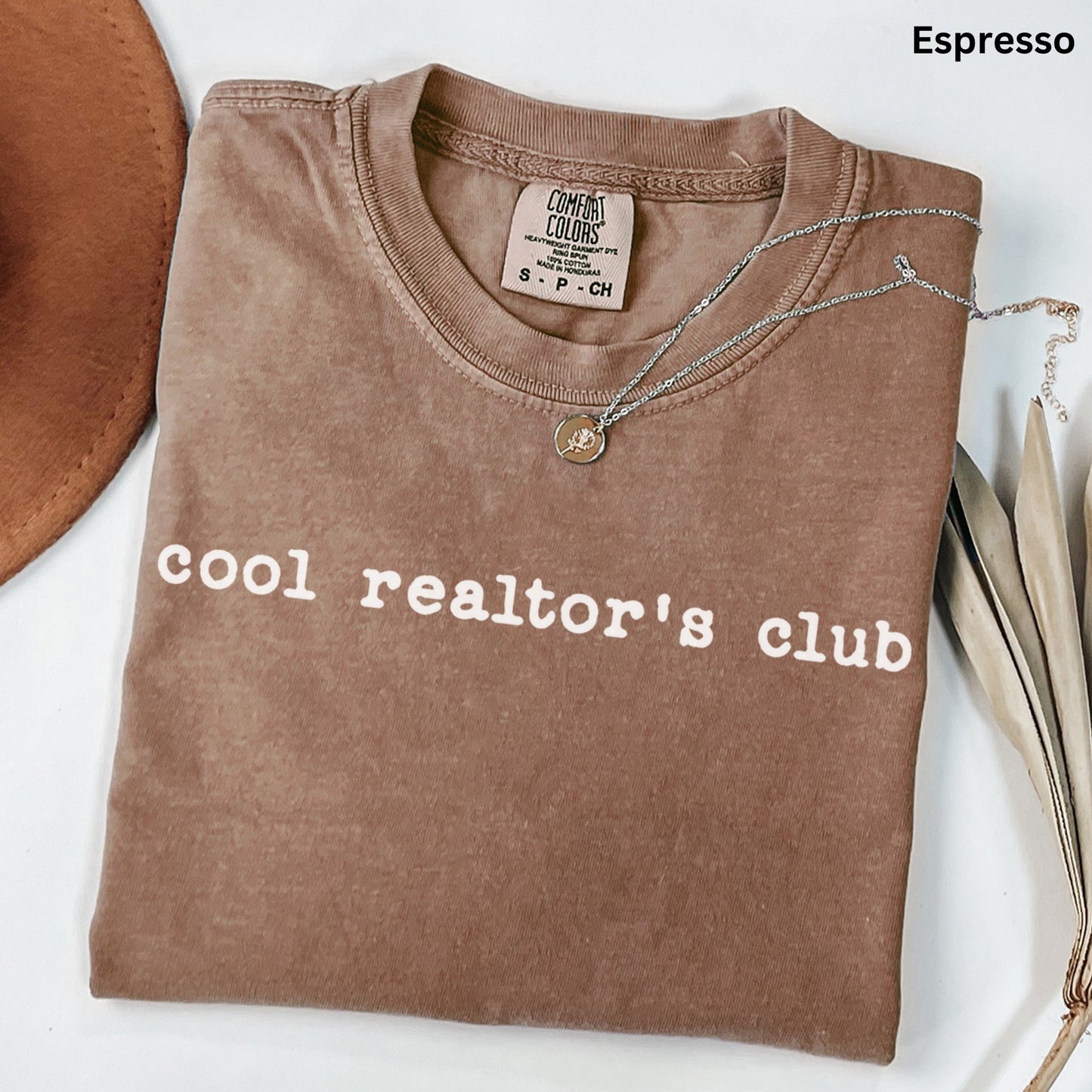 A brown t-shirt with the text "cool realtor's club" printed on it, along with a necklace and some leaves.