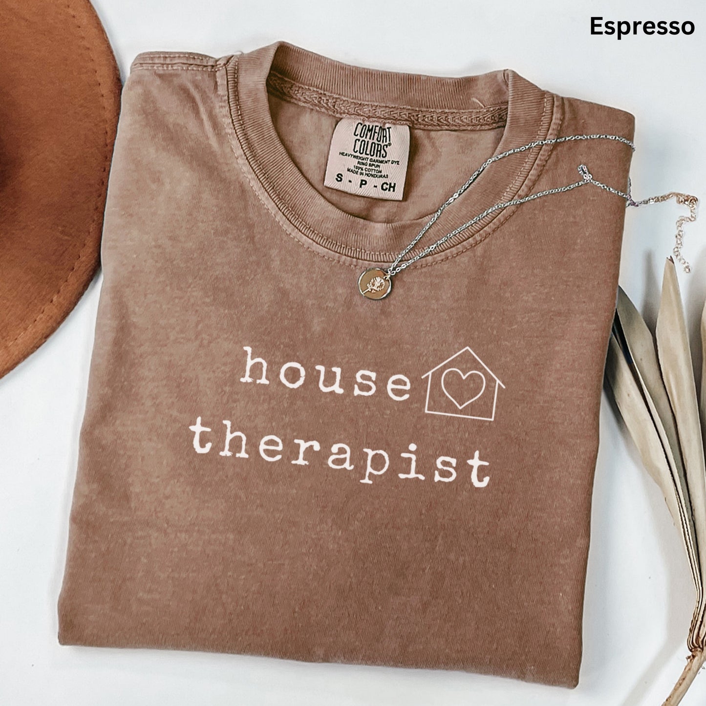a brown t-shirt with the text "house therapist" printed on it, along with a heart symbol and a house icon. The t-shirt is folded and placed on a white surface, with a brown hat and some leaves visible in the background.