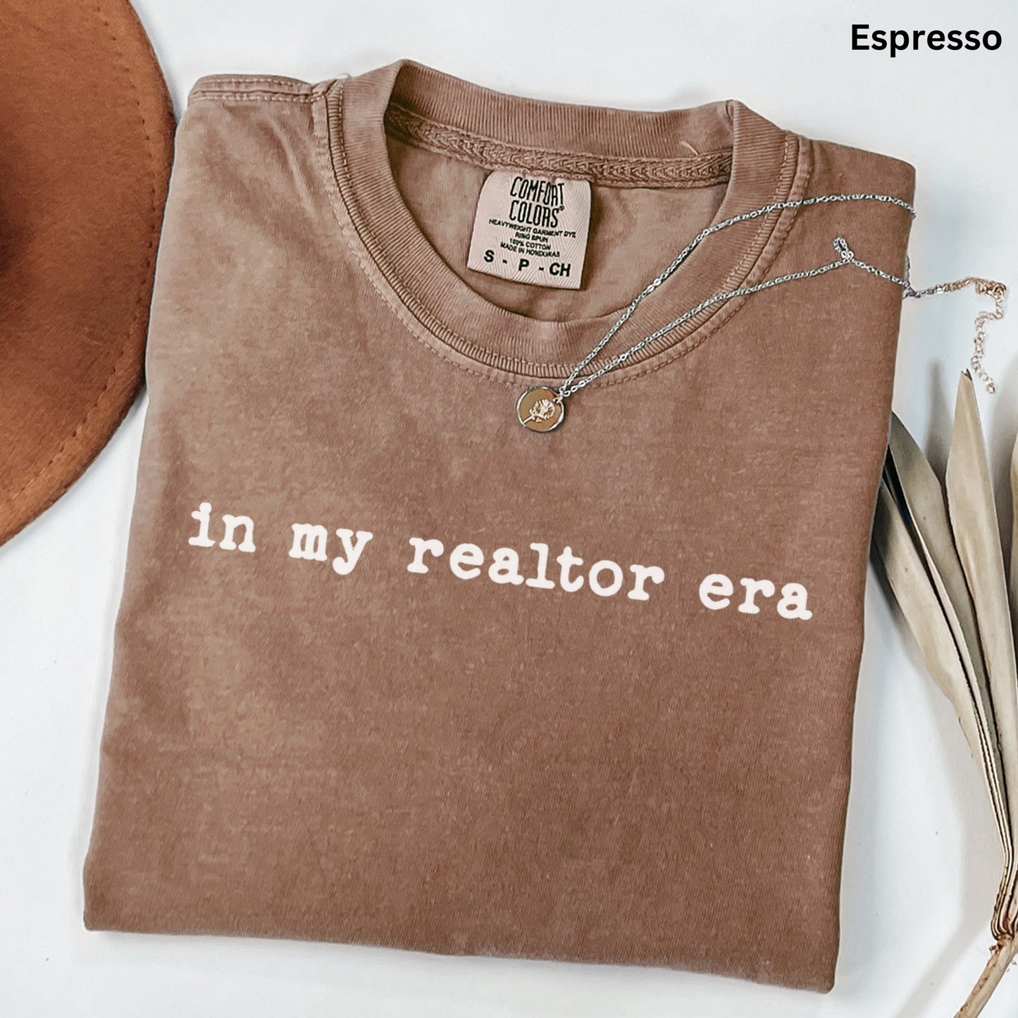 A brown t-shirt with the text "in my realtor era" printed on it, along with a necklace and some leaves.