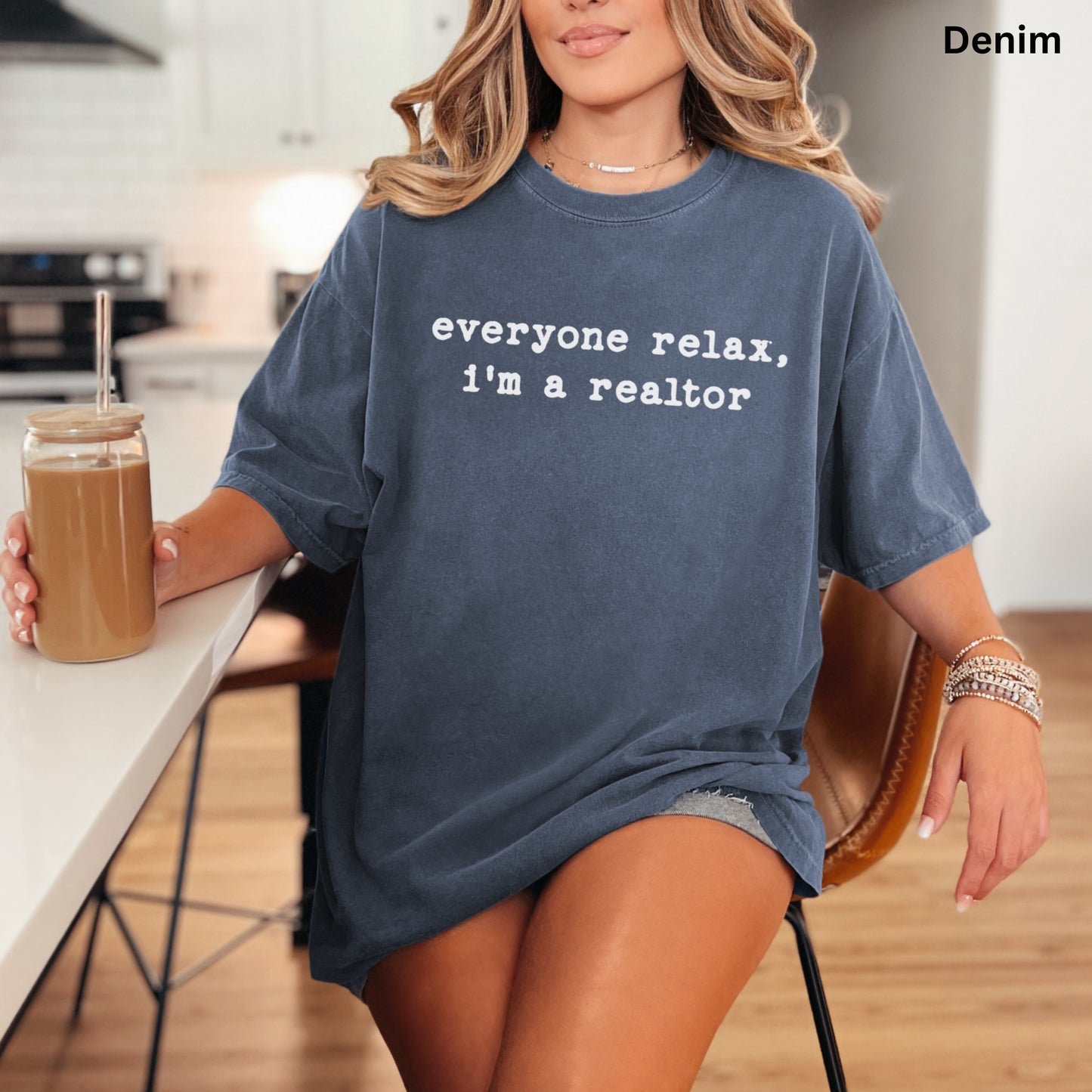 A woman wearing a blue t-shirt with the text "Everyone relax, I'm a realtor" is sitting at a kitchen counter, holding a drink.