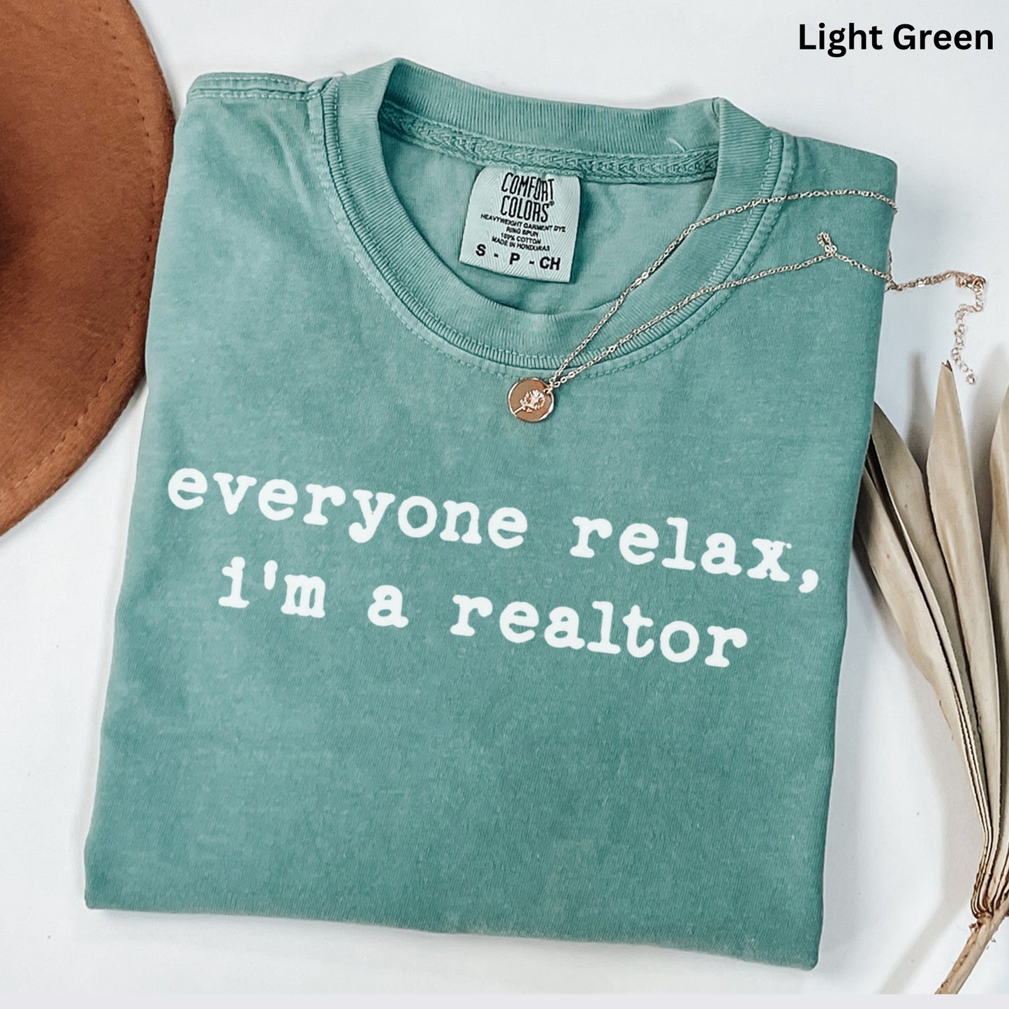A light green t-shirt with the text "Everyone relax, I'm a realtor" printed on it, along with a small gold pendant. The t-shirt is laid out on a white surface, and there is a brown hat and some dried leaves nearby.