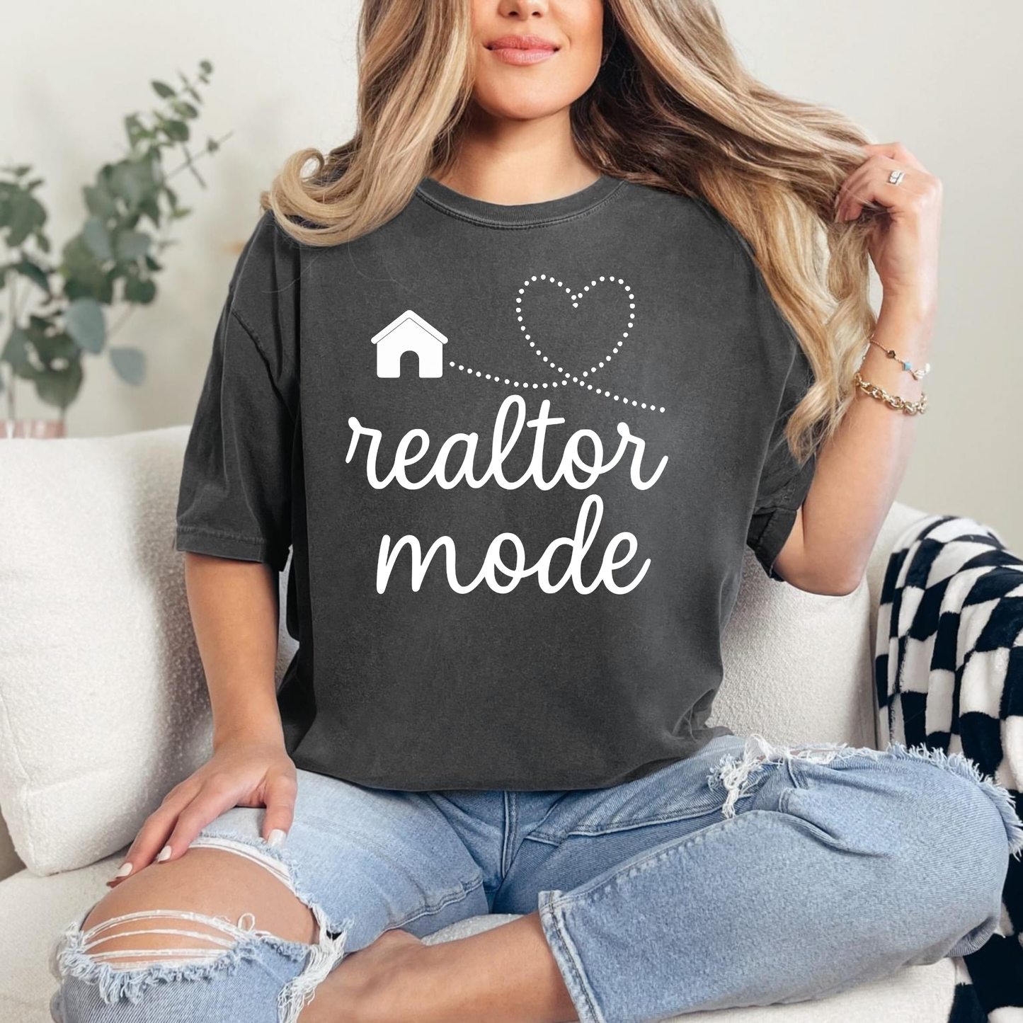 A woman sitting on a couch wearing a black t-shirt with the text "realtor mode" and a heart symbol, with her hair blowing in the wind.