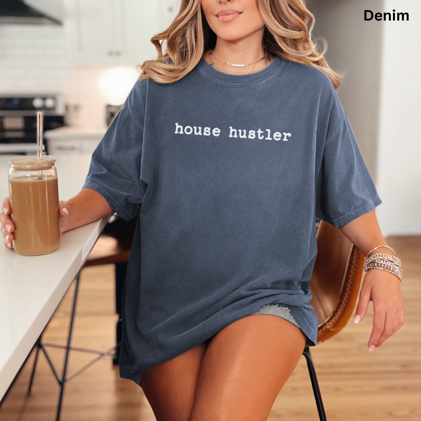 A woman wearing a blue t-shirt with the text "house hustler" is sitting at a kitchen counter, holding a drink in a glass.