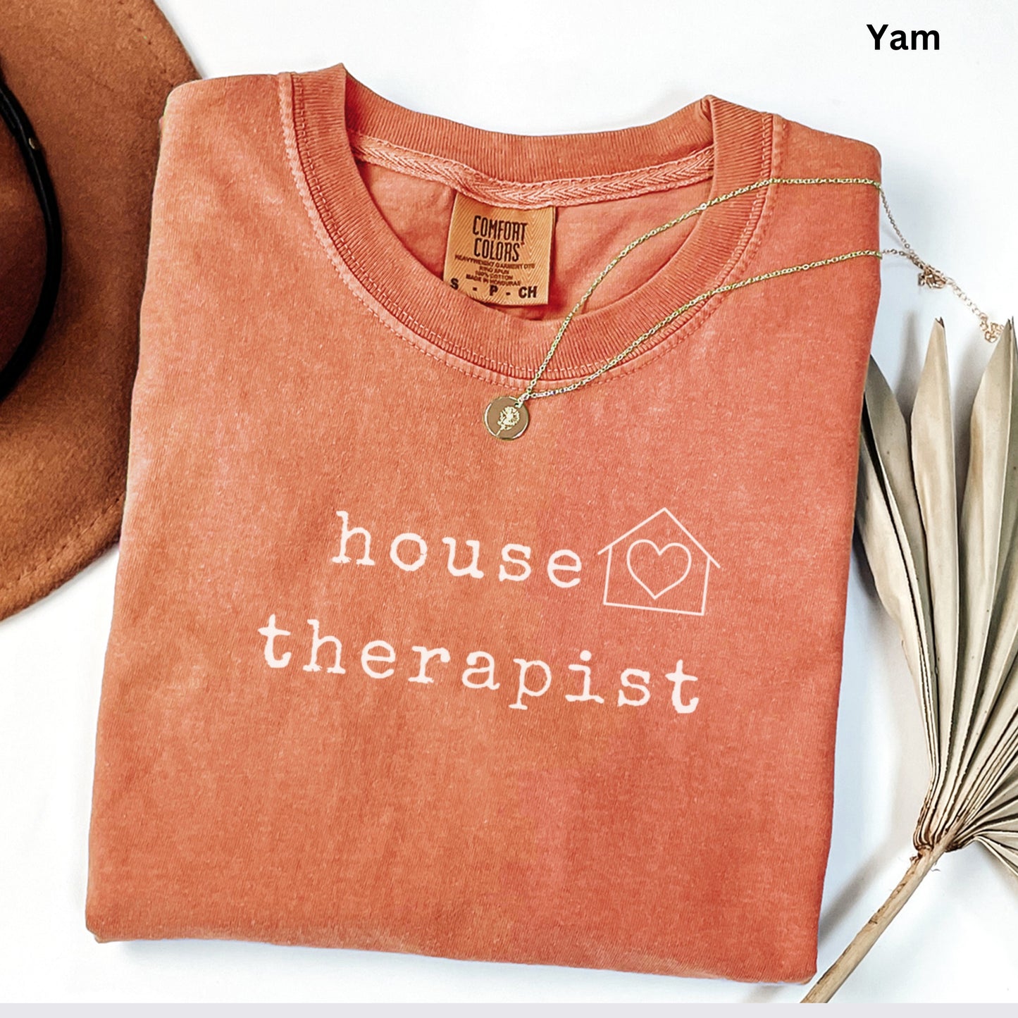 a red t-shirt with the text "house therapist" printed on it, along with a small house symbol. The t-shirt is folded and placed on a white surface, with a brown hat and some leaves visible in the background.