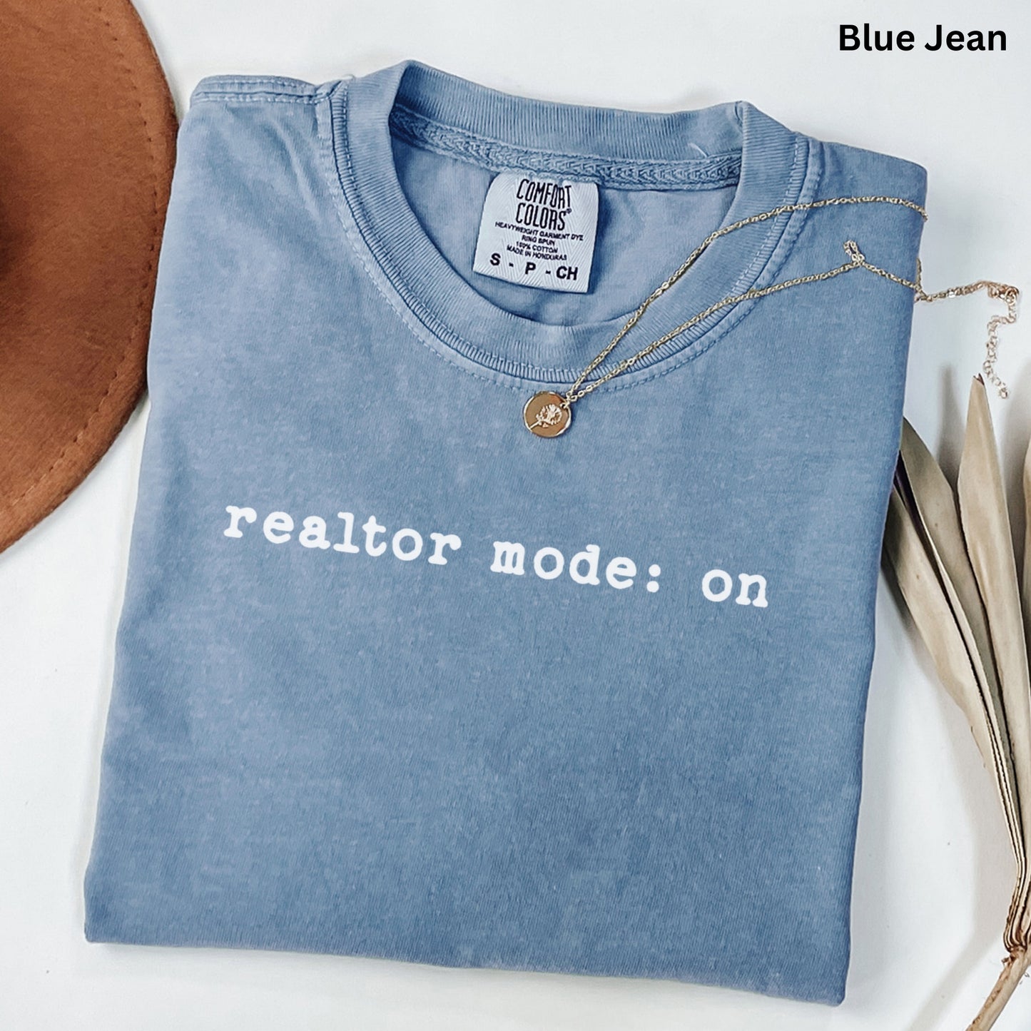 A blue t-shirt with the text "realtor mode: on" printed on it, along with a necklace and some leaves.
