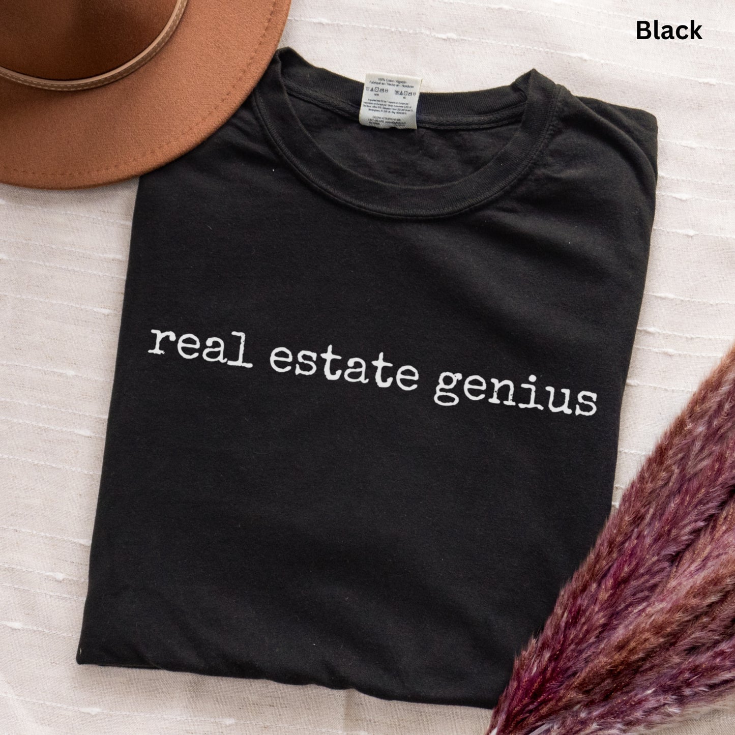 a black t-shirt with the text "real estate Genius" printed on it, along with a brown hat and some purple flowers.