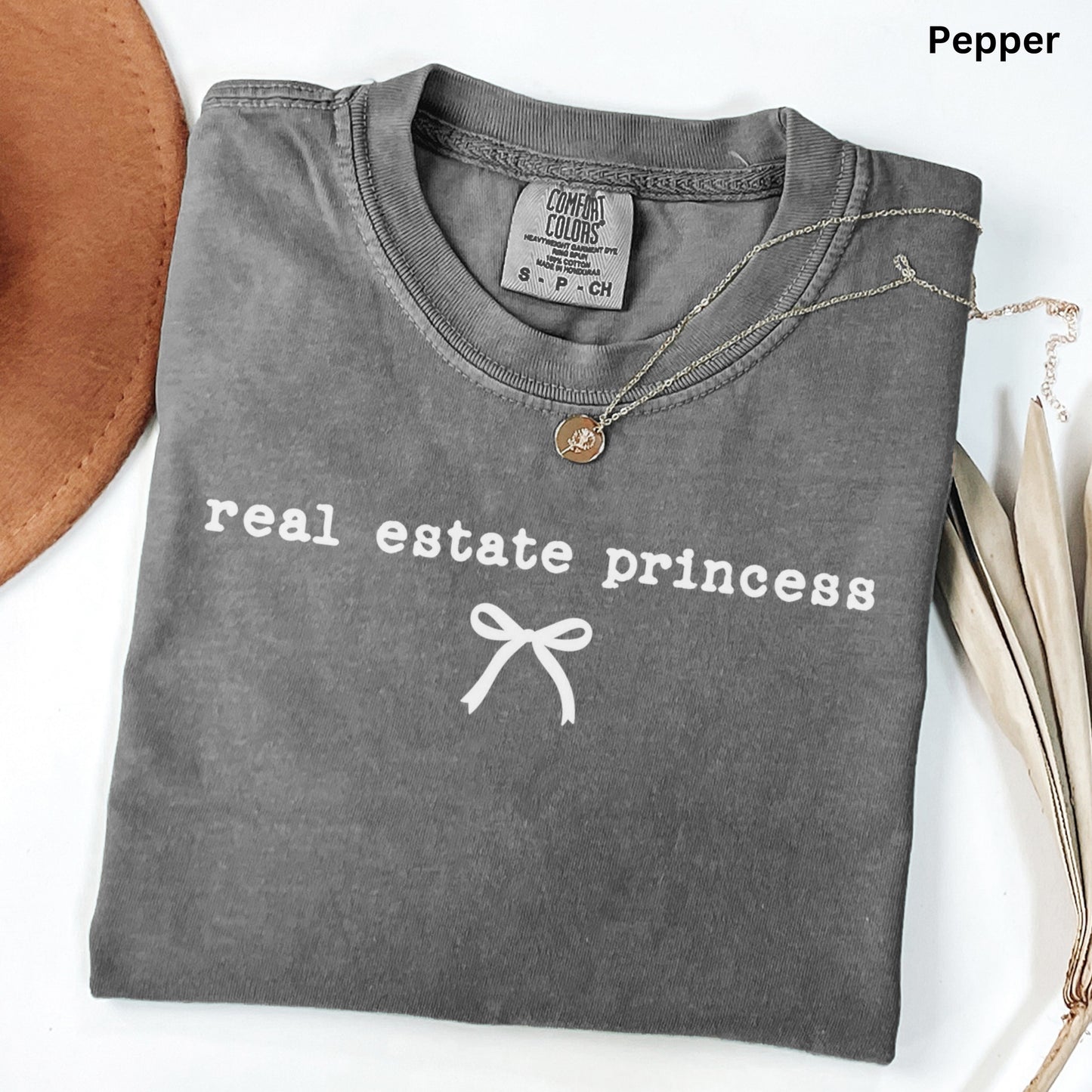 a gray t-shirt with the text "real estate princess" printed on it, along with a ribbon design. The t-shirt is folded and placed on a white surface, with a brown hat and some leaves visible in the background.