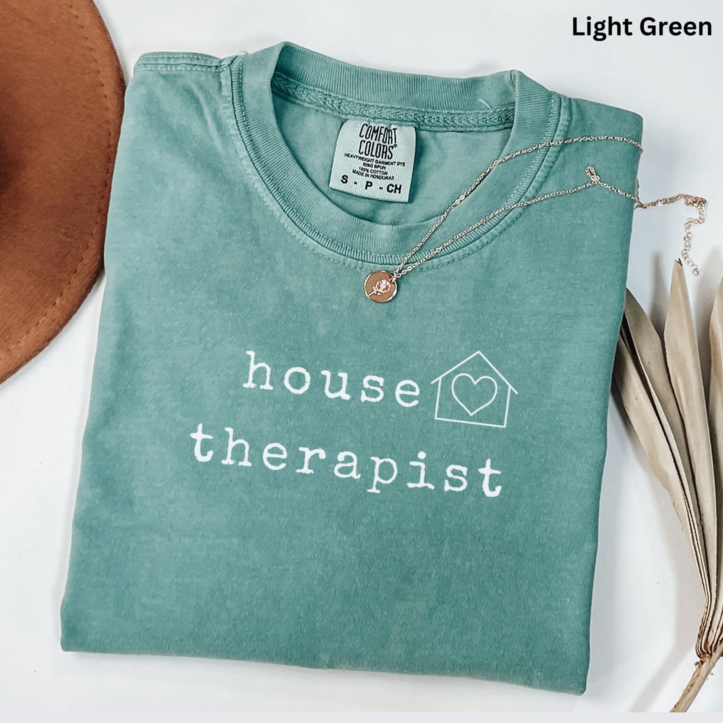 A light green t-shirt with the text "house therapist" printed on it, along with a heart symbol and a house icon. The t-shirt is folded and placed on a white surface, with a brown hat and some dried leaves nearby.