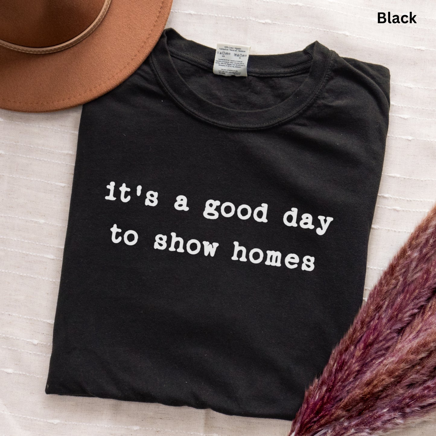 A black t-shirt with white text that reads "It's a good day to show homes" is displayed on a white surface, accompanied by a brown hat and a purple plant.