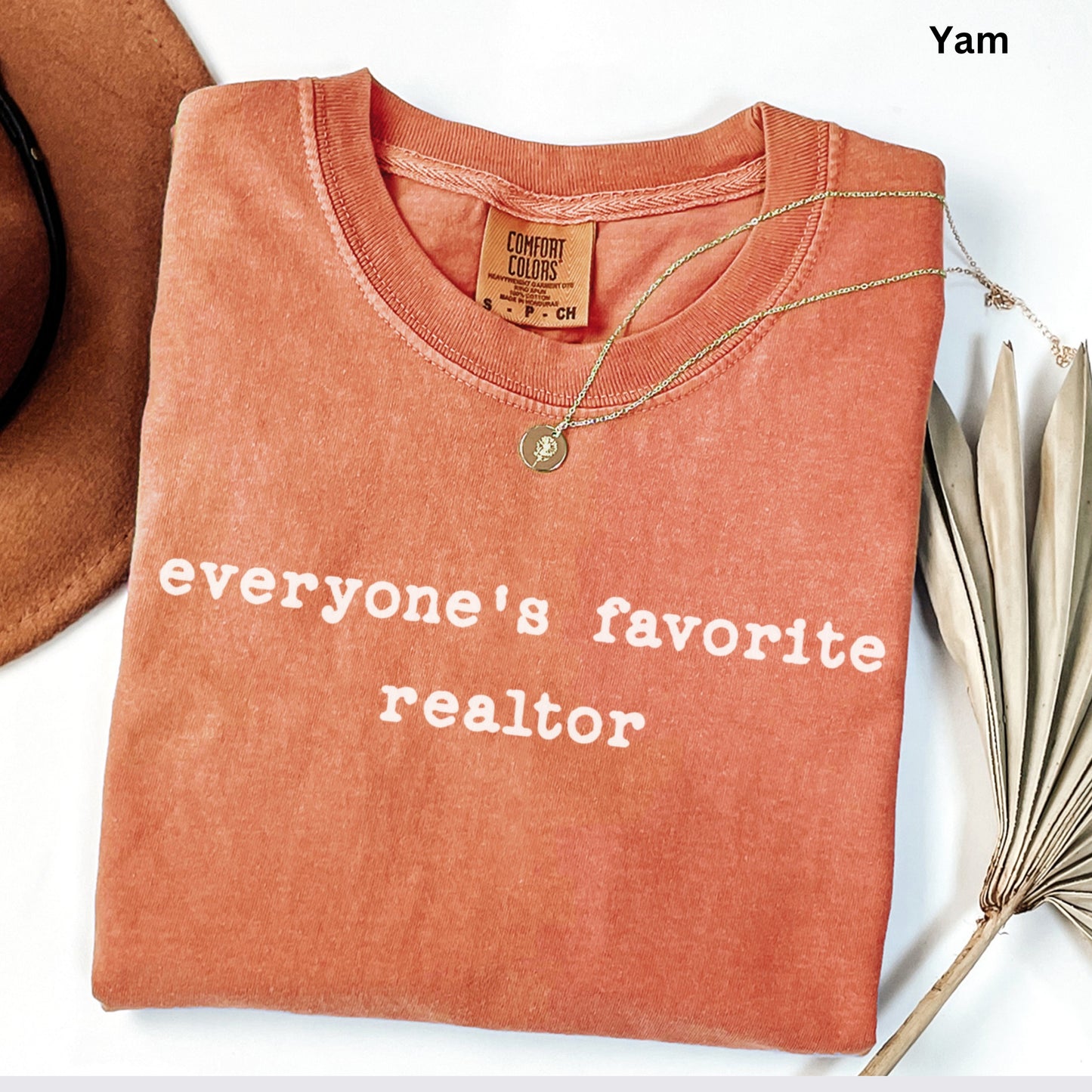 a red t-shirt with the text "Everyone's favorite realtor" printed on it, along with a gold necklace and a brown hat.
