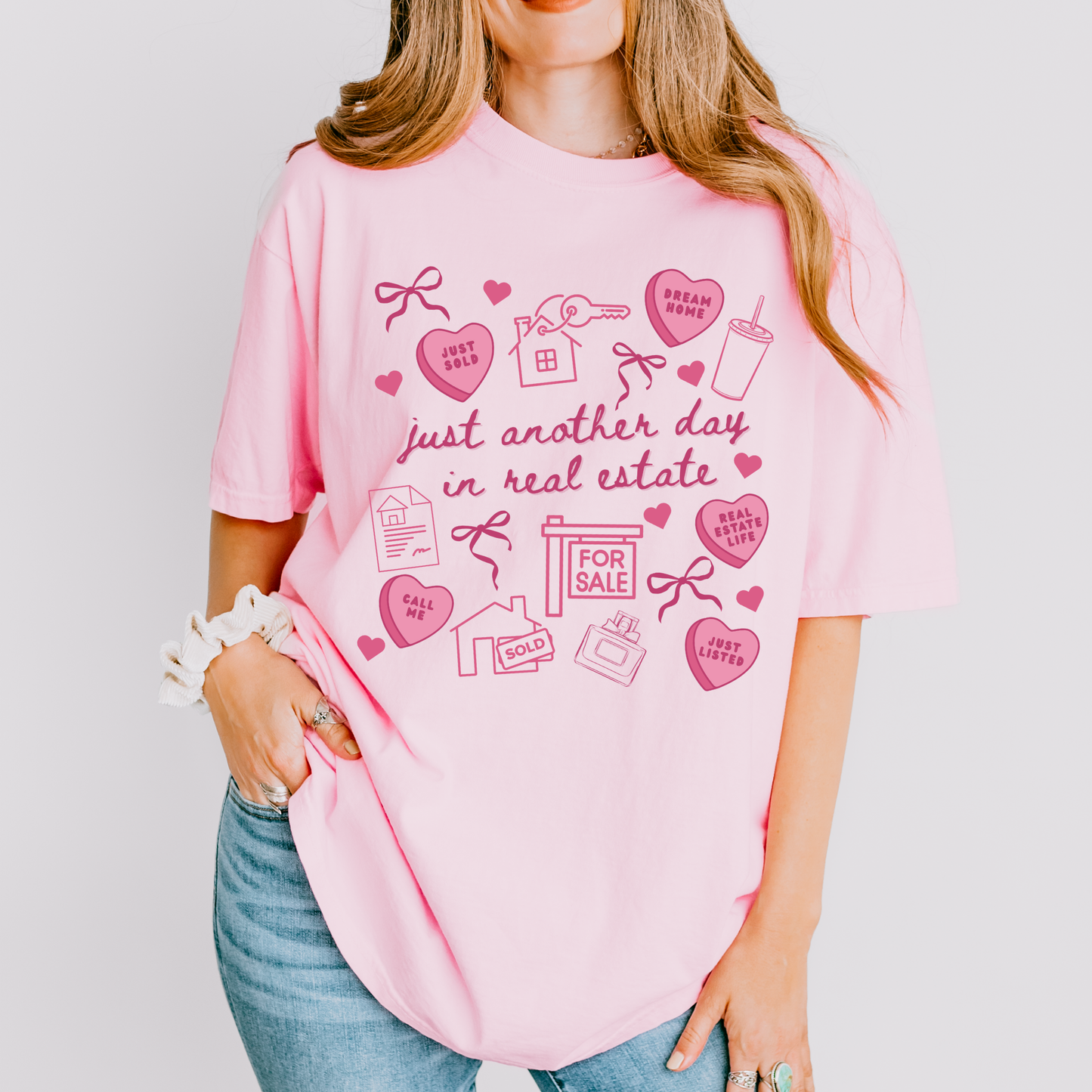 A woman wearing a pink t-shirt with a graphic design of various real estate-related icons and text, including a house, keys, and a "For Sale" sign.