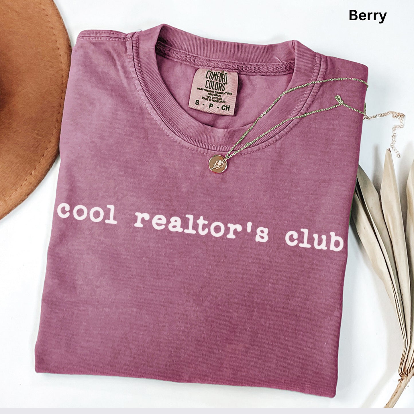 A pink t-shirt with the text "cool realtor's club" printed on it, along with a necklace and some leaves.