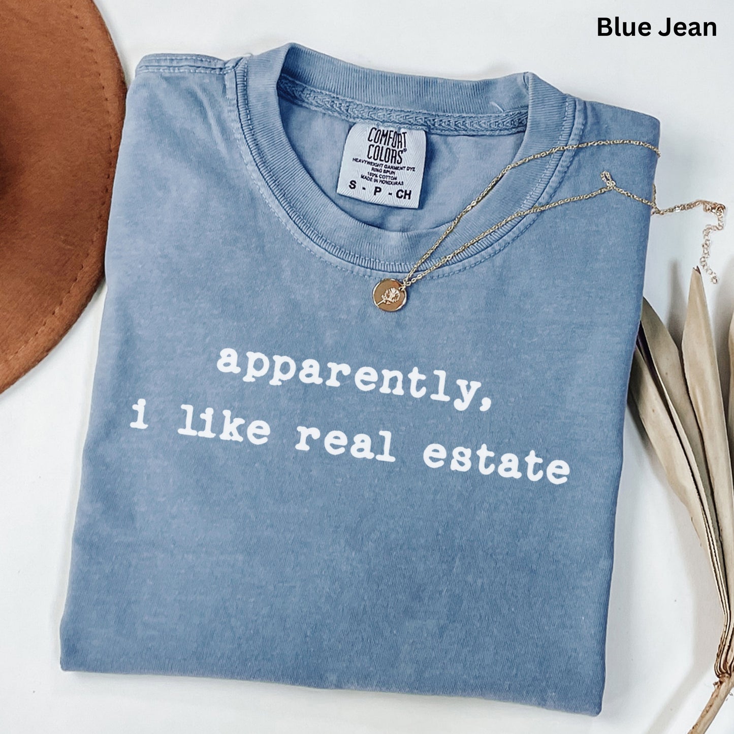 A blue t-shirt with the text "apparently, I like real estate" printed on it, along with a small gold medallion. The t-shirt is laid out on a white surface, and there is a brown hat and some leaves nearby.