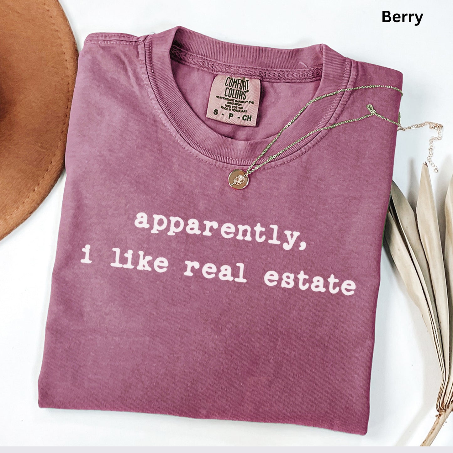 A purple t-shirt with the text "apparently, I like real estate" printed on it, along with a small gold pendant. The t-shirt is folded and placed on a white surface, with a brown hat and some leaves visible in the background.