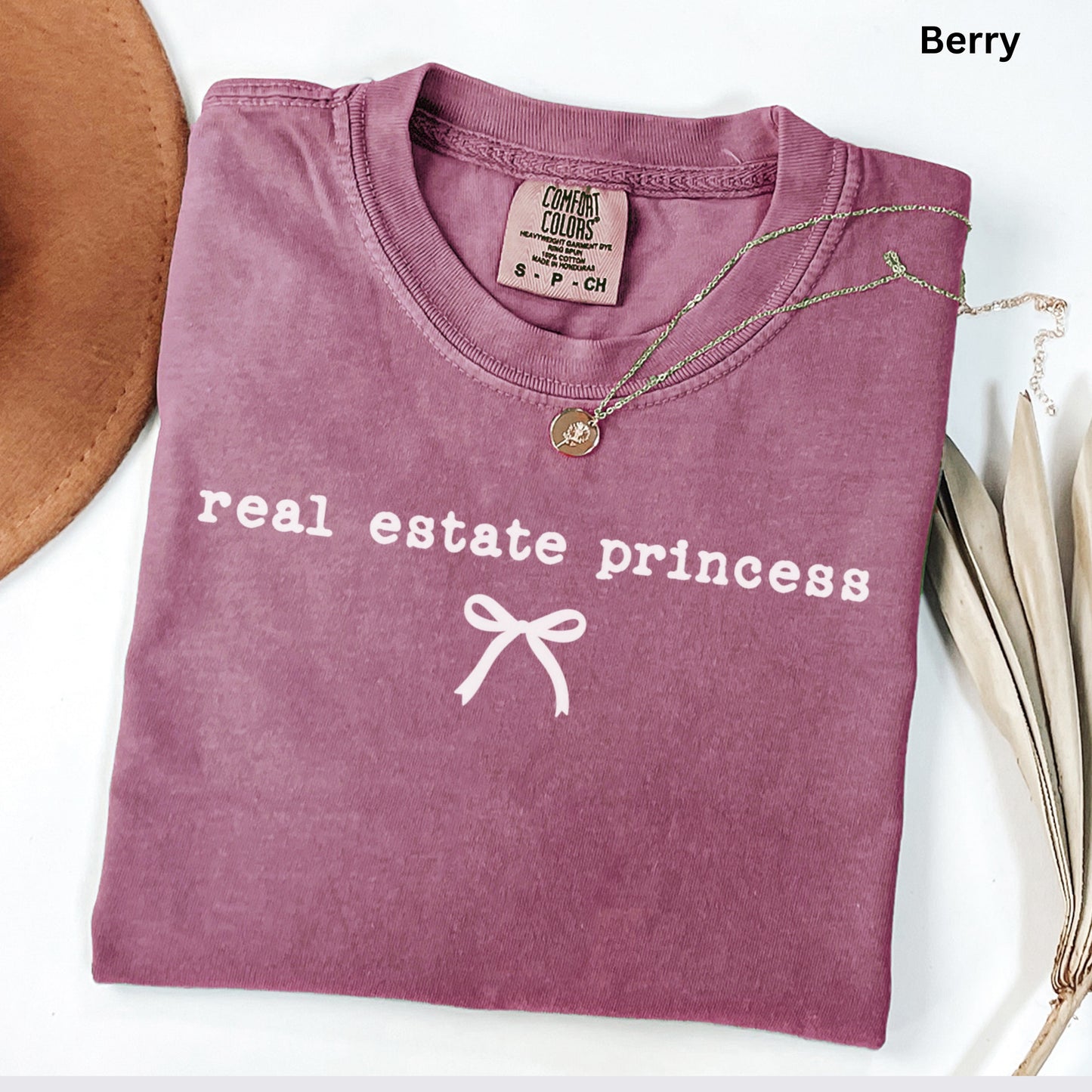 A pink t-shirt with the text "real estate princess" printed on it, along with a small white bow symbol. The t-shirt is folded and placed on a white surface, accompanied by a brown hat and some dried leaves.