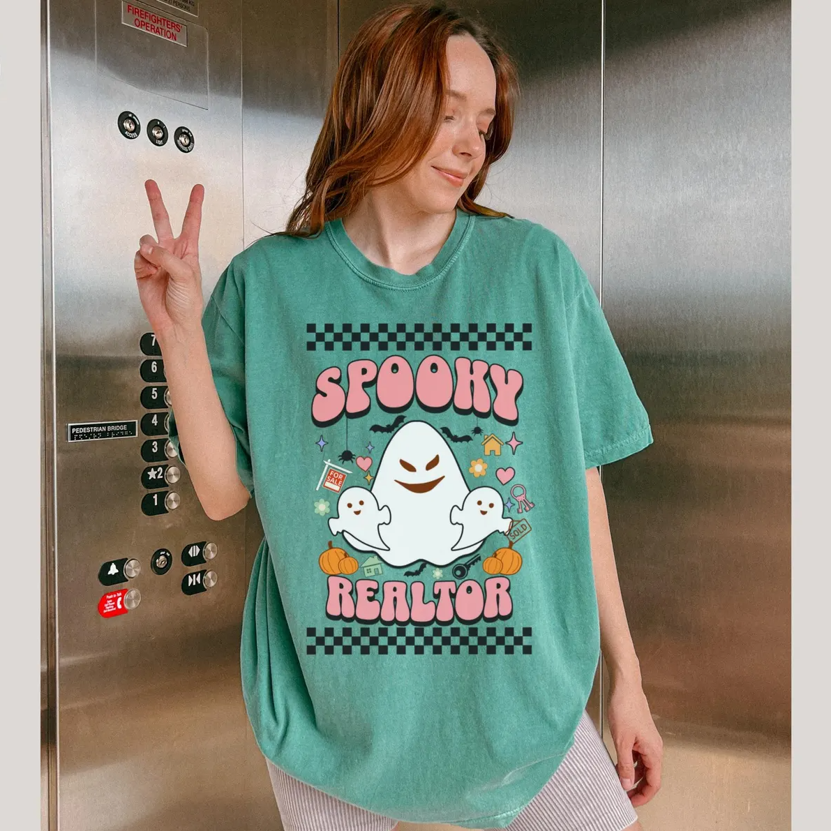 A woman wearing a green t-shirt with a graphic design of a ghost and the text "SPOKEY REALTOR" is posing in front of an elevator.