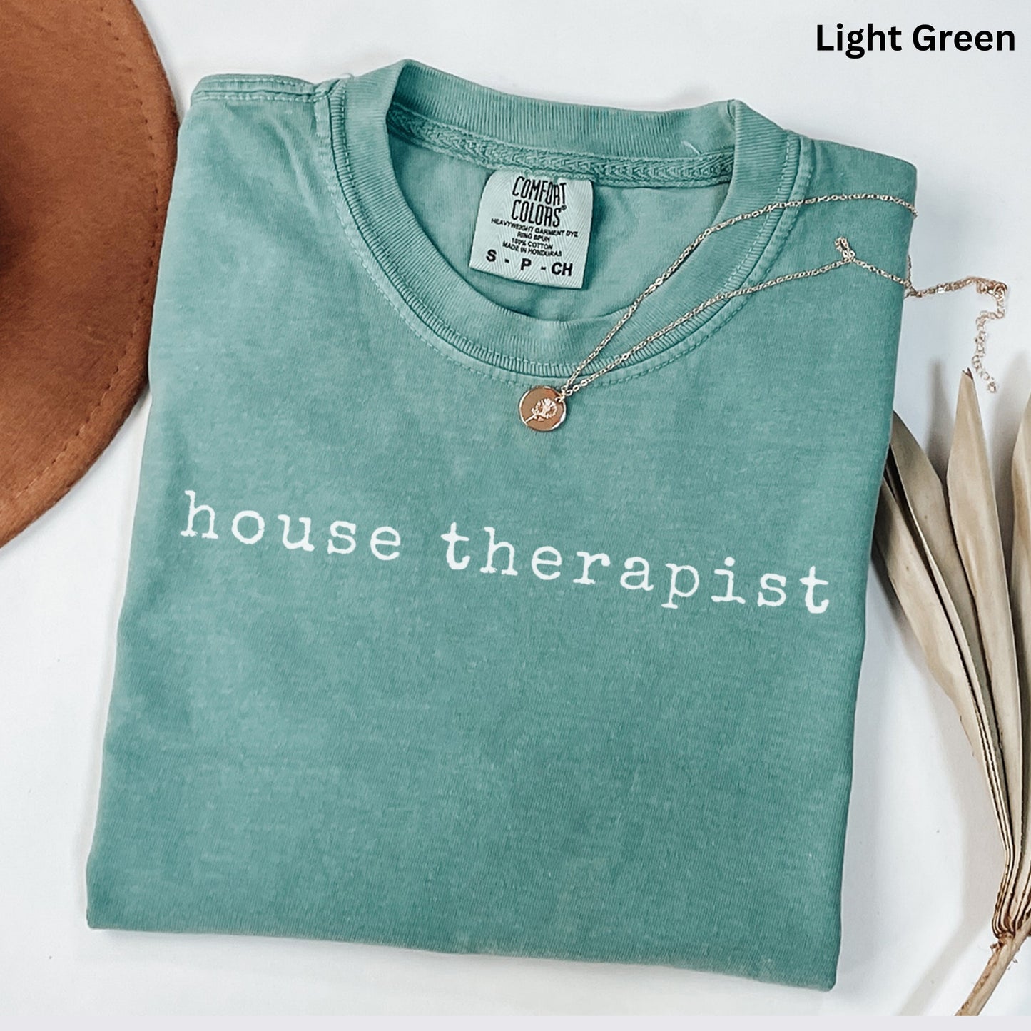A light green t-shirt with the text "house therapist" printed on it, along with a necklace and a brown hat.