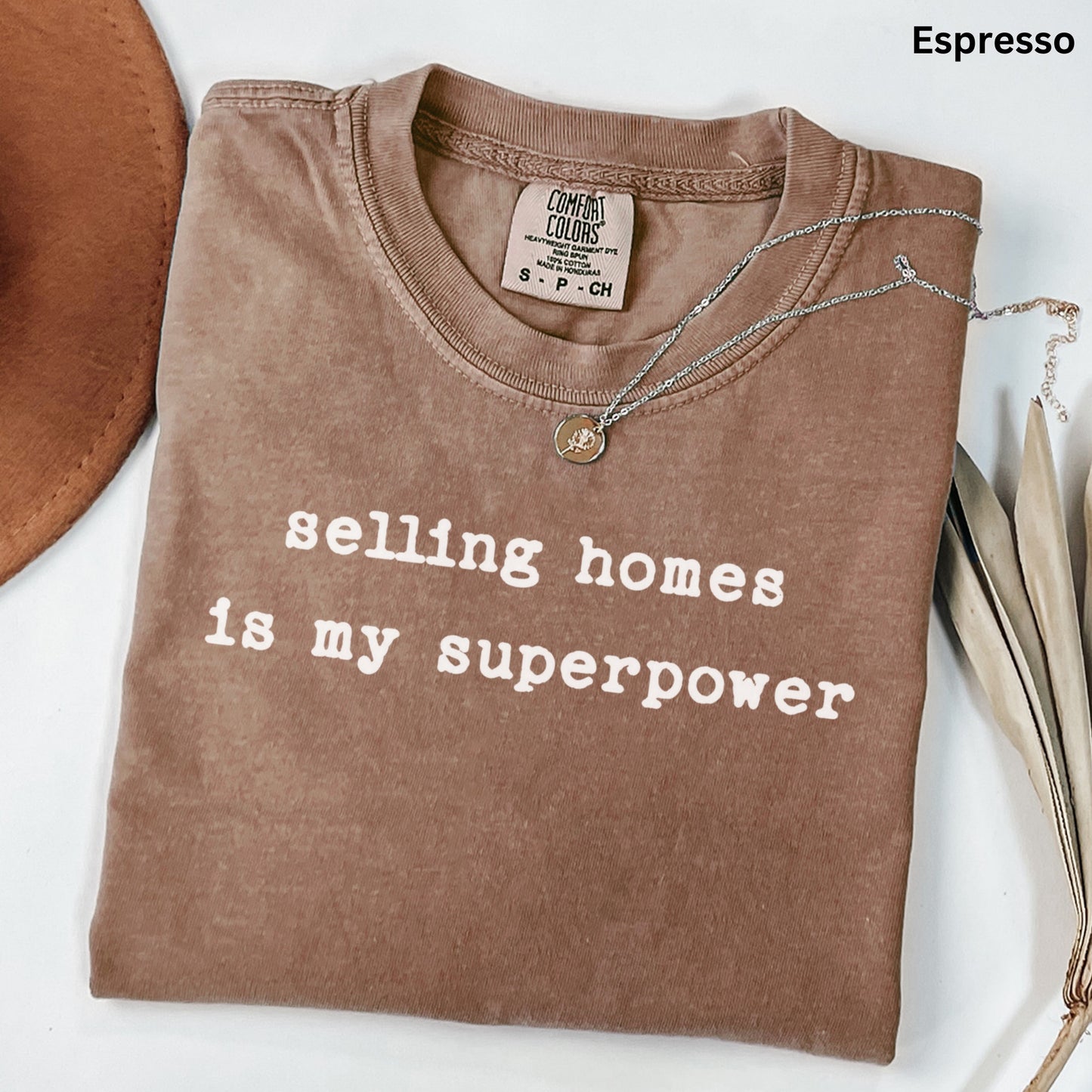 A brown t-shirt with the text "selling homes is my superpower" printed on it, along with a necklace and some leaves.