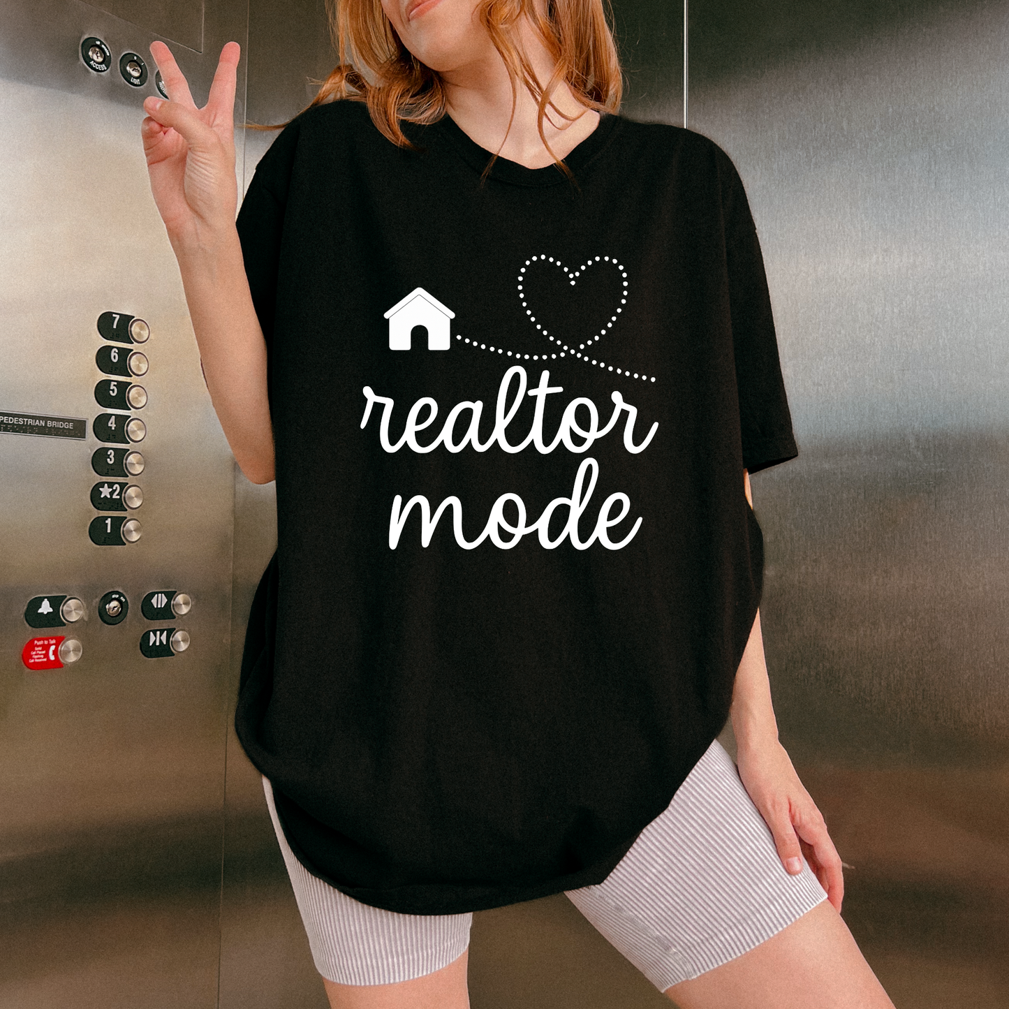 A person wearing a black t-shirt with the text "realtor mode" and a heart symbol, standing in front of an elevator.