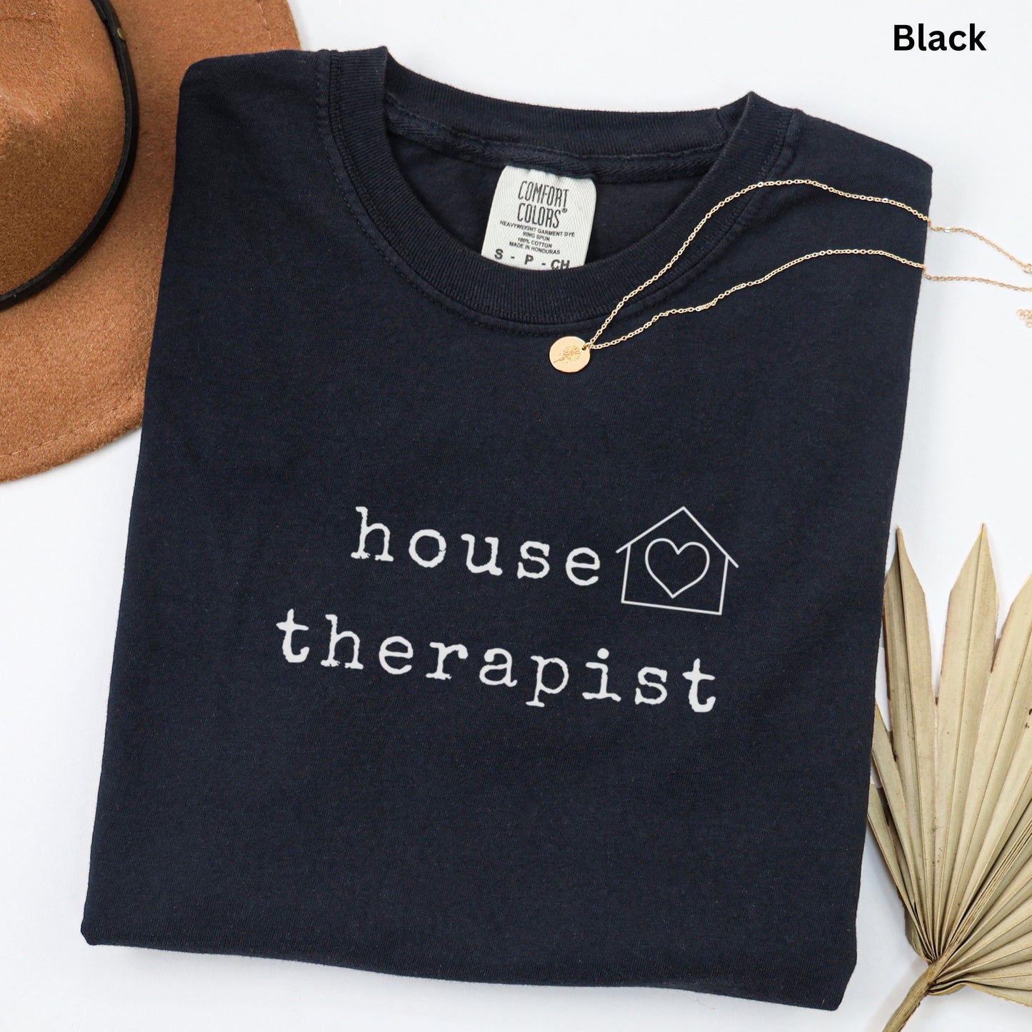 A black t-shirt with the text "house therapist" printed on it, along with a heart symbol and a house icon. The t-shirt is laid out on a white surface, with a brown hat and a leaf visible in the background.