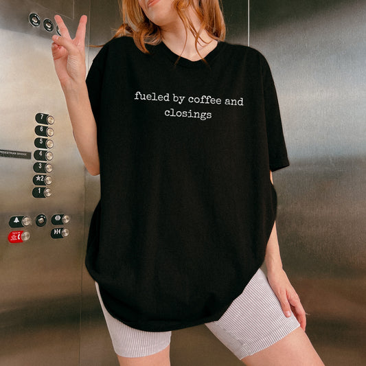 A person wearing a black t-shirt with the text "fueled by coffee and closings" printed on it, standing in front of an elevator control panel.