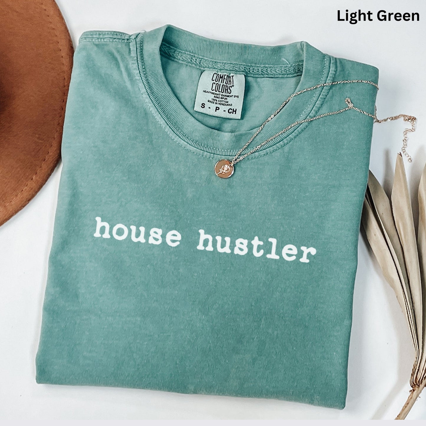 A light green t-shirt with the text "house hustler" printed on it, along with a necklace and a brown hat.