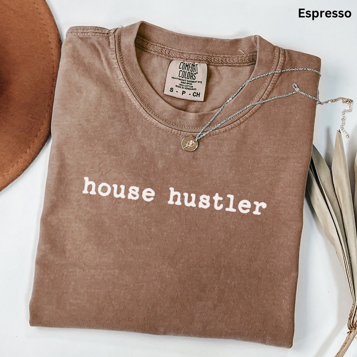 a brown t-shirt with the text "house hustler" printed on it, along with a necklace and some leaves.