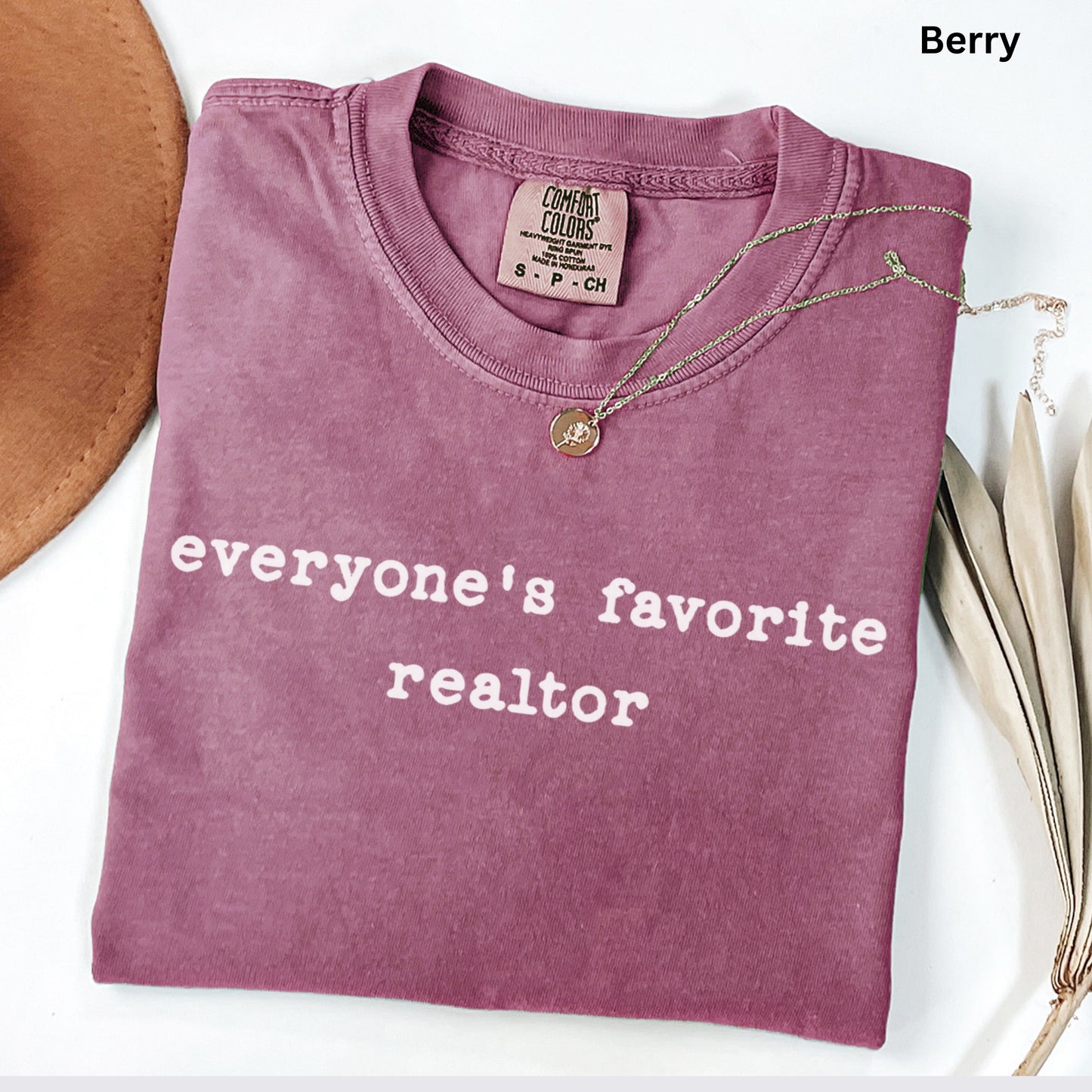 A pink t-shirt with the text "Everyone's favorite realtor" printed on it, along with a small gold pendant. The t-shirt is folded and placed on a white surface, with a brown hat and some leaves visible in the background.
