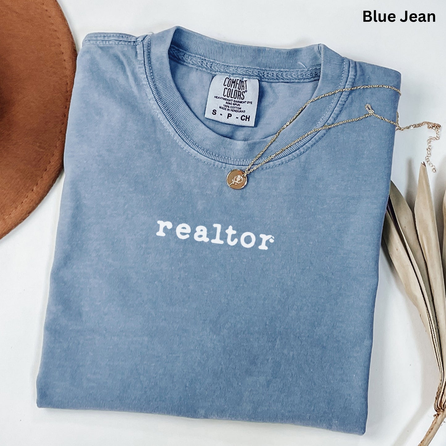 A blue t-shirt with the word "realtor" printed on it, along with a necklace and some leaves, is displayed on a white surface.
