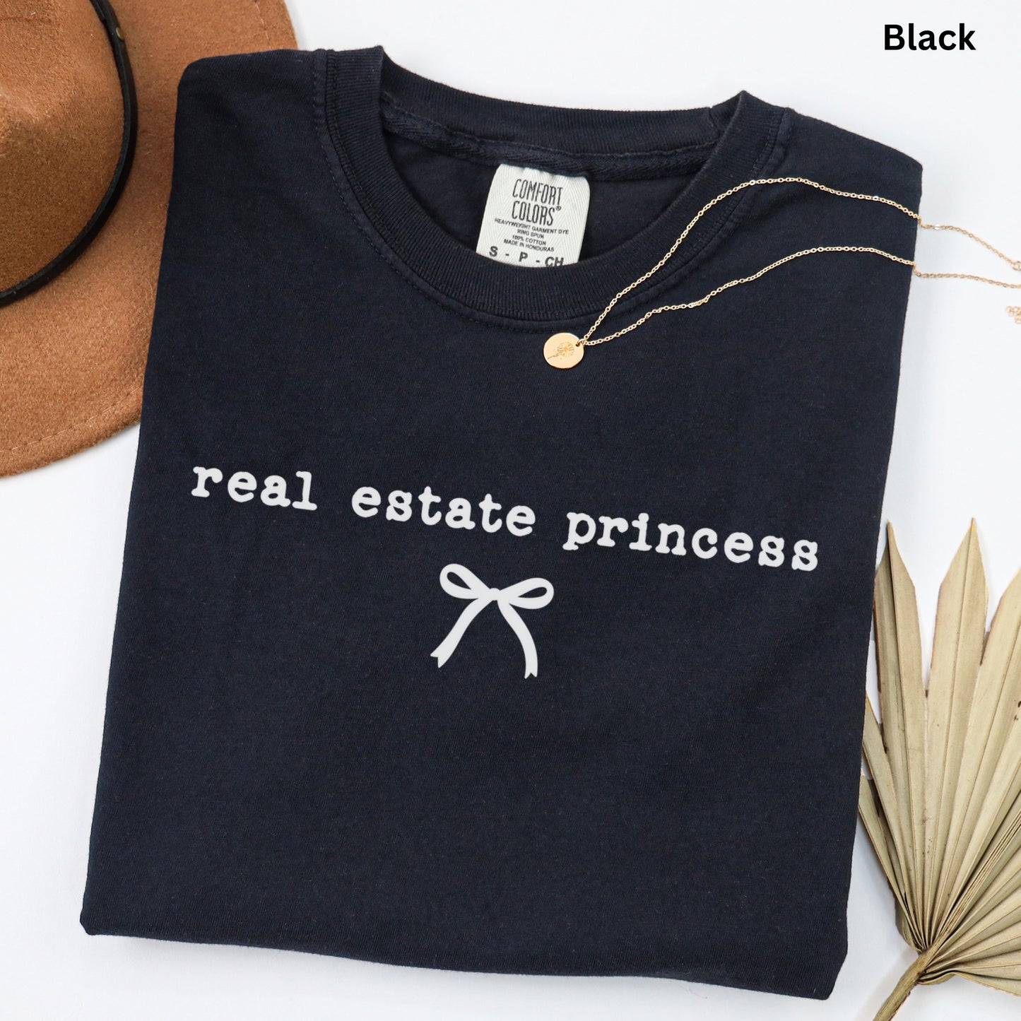 a black t-shirt with the text "real estate princess" printed on it, along with a ribbon design. The t-shirt is laid out on a white surface, and there is a brown hat and a leaf visible in the background.