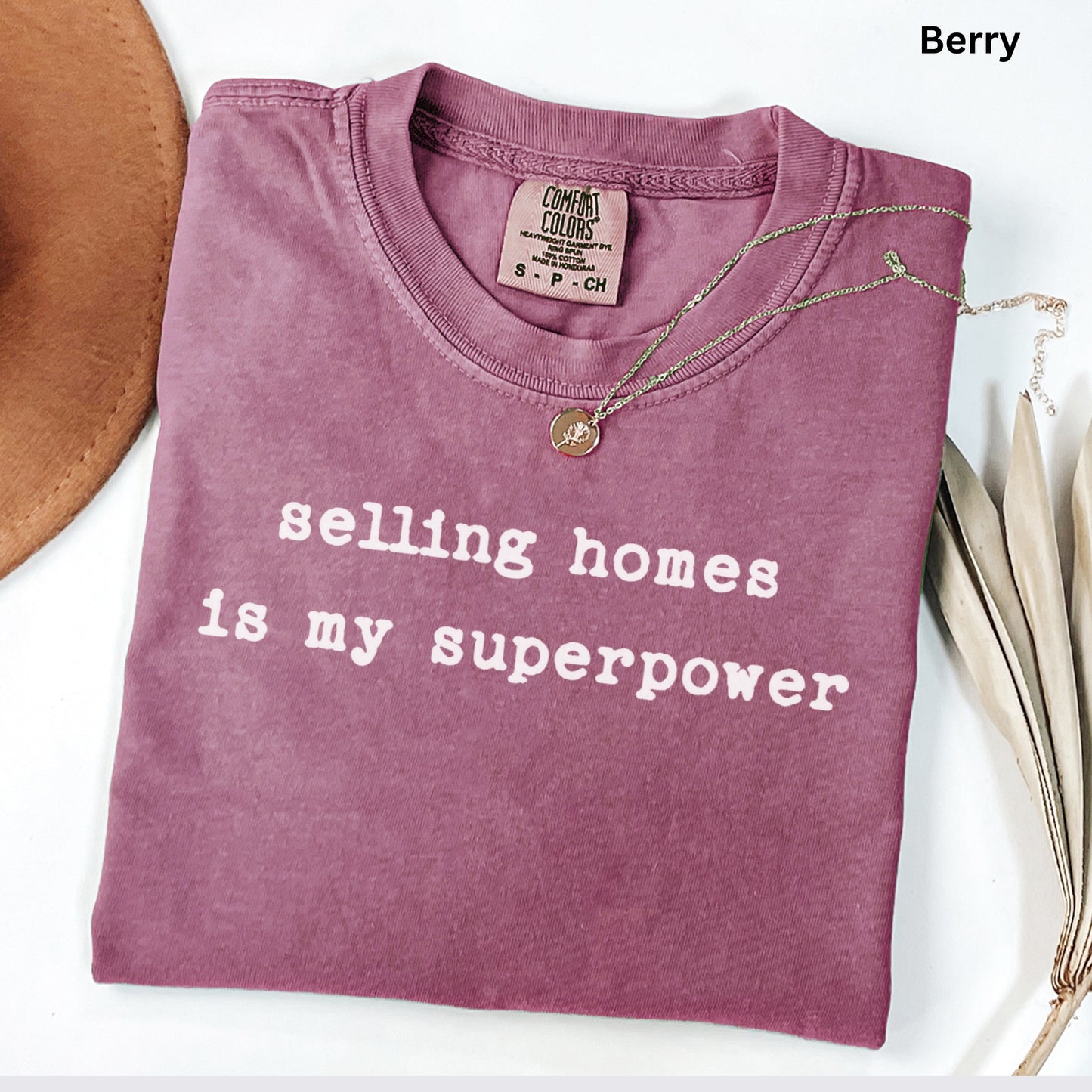 A purple t-shirt with the text "selling homes is my superpower" printed on it, along with a necklace and a hat.