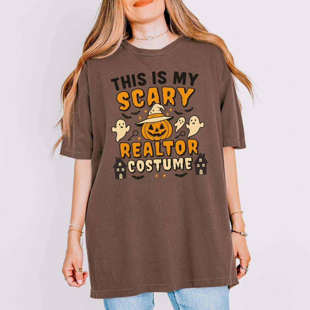 A woman wearing a brown t-shirt with a Halloween-themed design, including a jack-o-lantern, ghost, and houses.