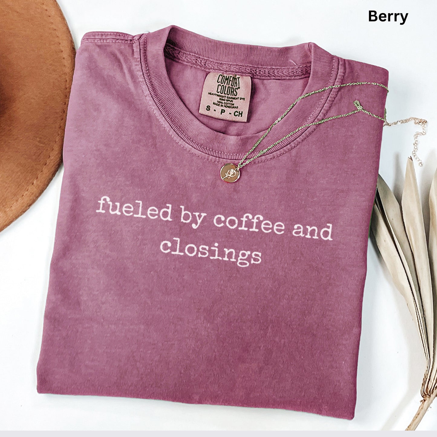 A pink t-shirt with the text "fused by coffee and closings" printed on it, along with a necklace and a brown hat.