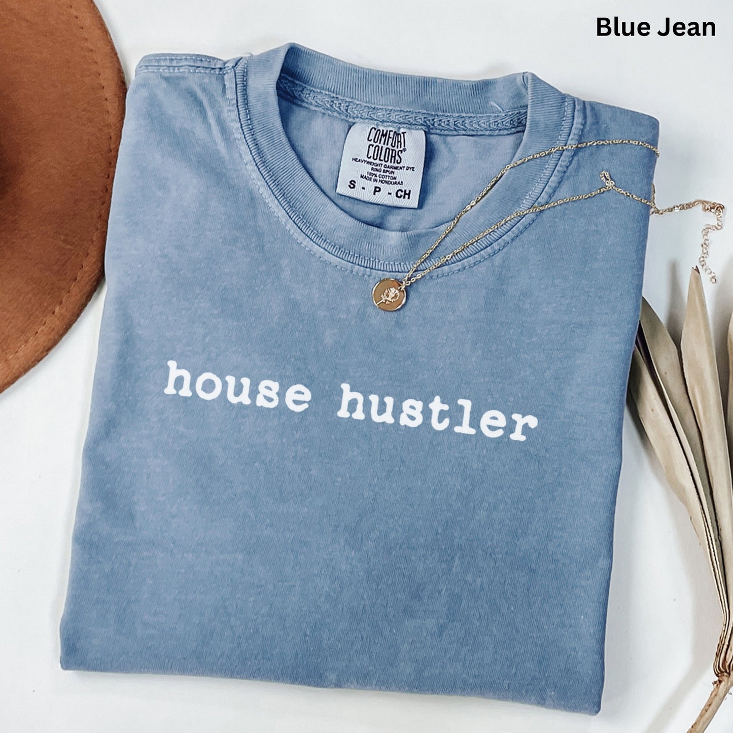 A blue t-shirt with the text "house hustler" printed on it, along with a necklace and some leaves.