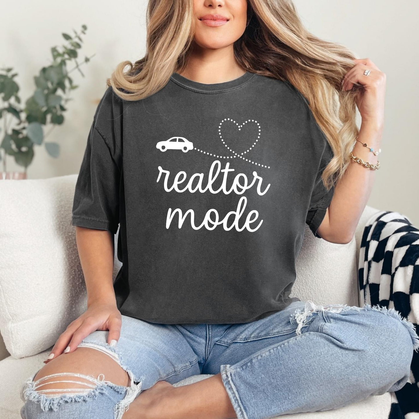 A woman sitting on a couch wearing a black t-shirt with the text "realtor mode" and a heart symbol, with her hair blowing in the wind.