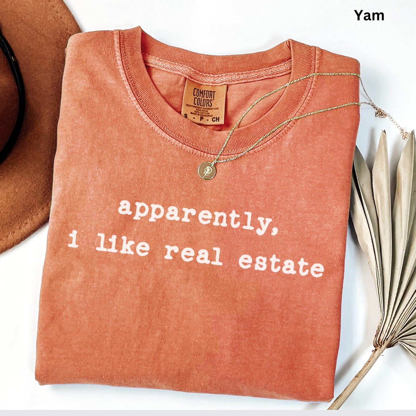a red t-shirt with the text "apparently, I like real estate" printed on it, along with a gold necklace and a brown hat.