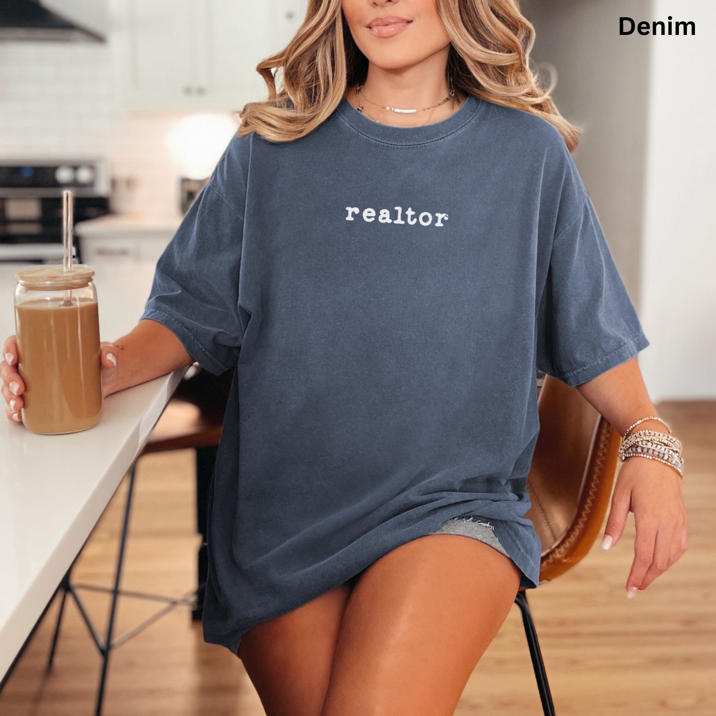 A woman wearing a blue t-shirt with the word "realator" printed on it, sitting at a kitchen counter with a drink in a glass.