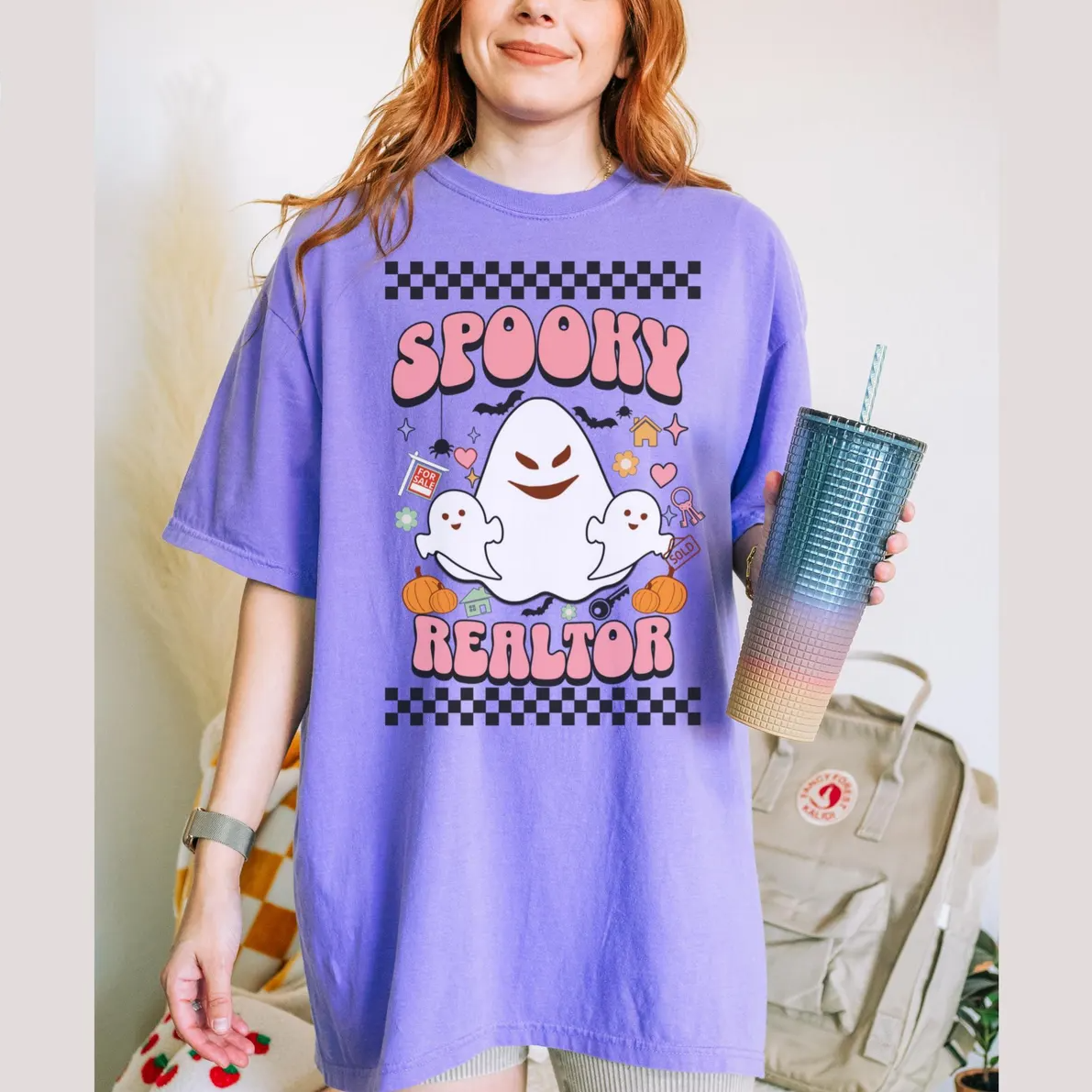 A woman wearing a purple t-shirt with a graphic design of a ghost and the text "SPOKEY REALTOR" is holding a cup with a straw.