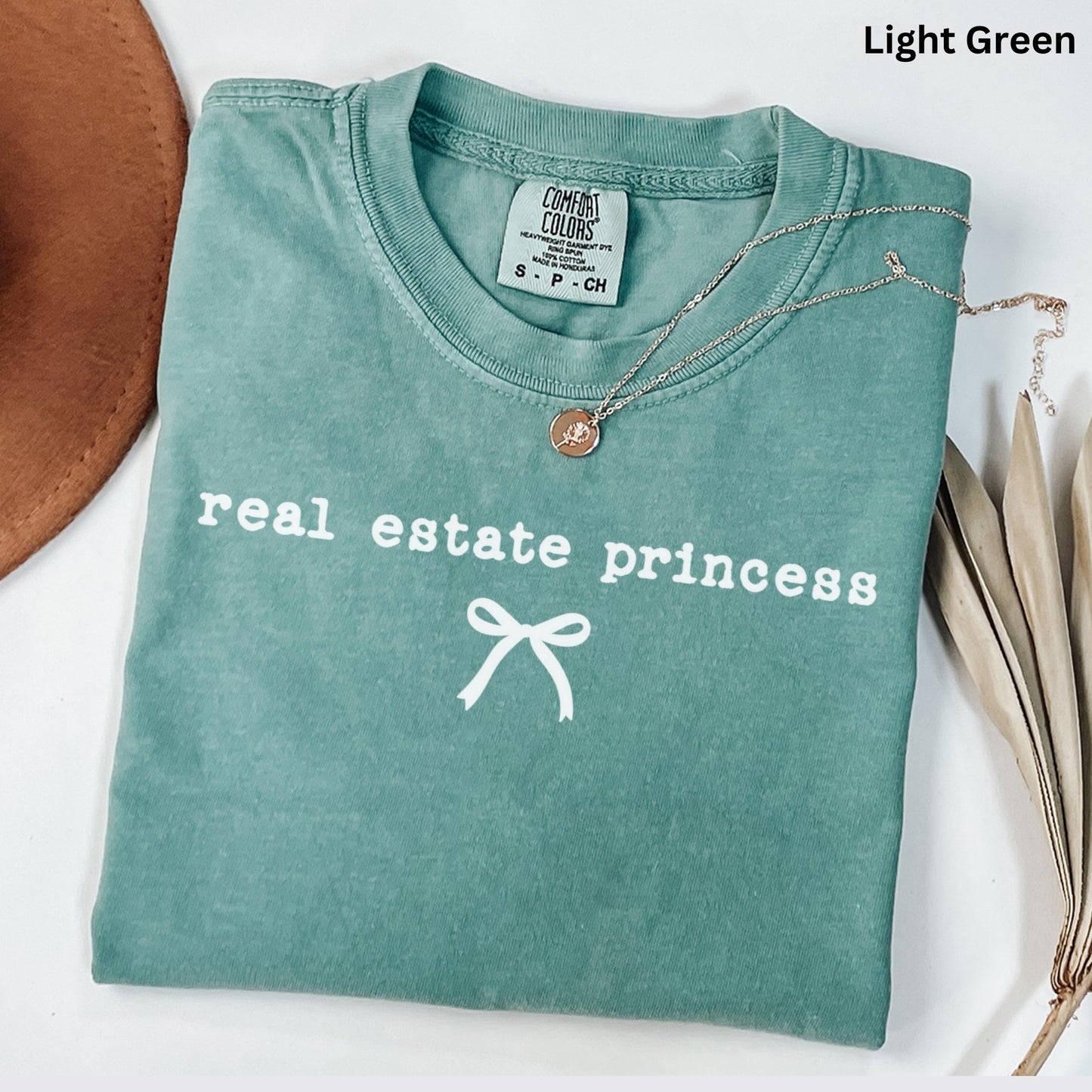 A light green t-shirt with the text "real estate princess" printed on it, along with a small white ribbon design. The t-shirt is folded and placed on a white surface, with a brown hat and some dried leaves visible in the background.