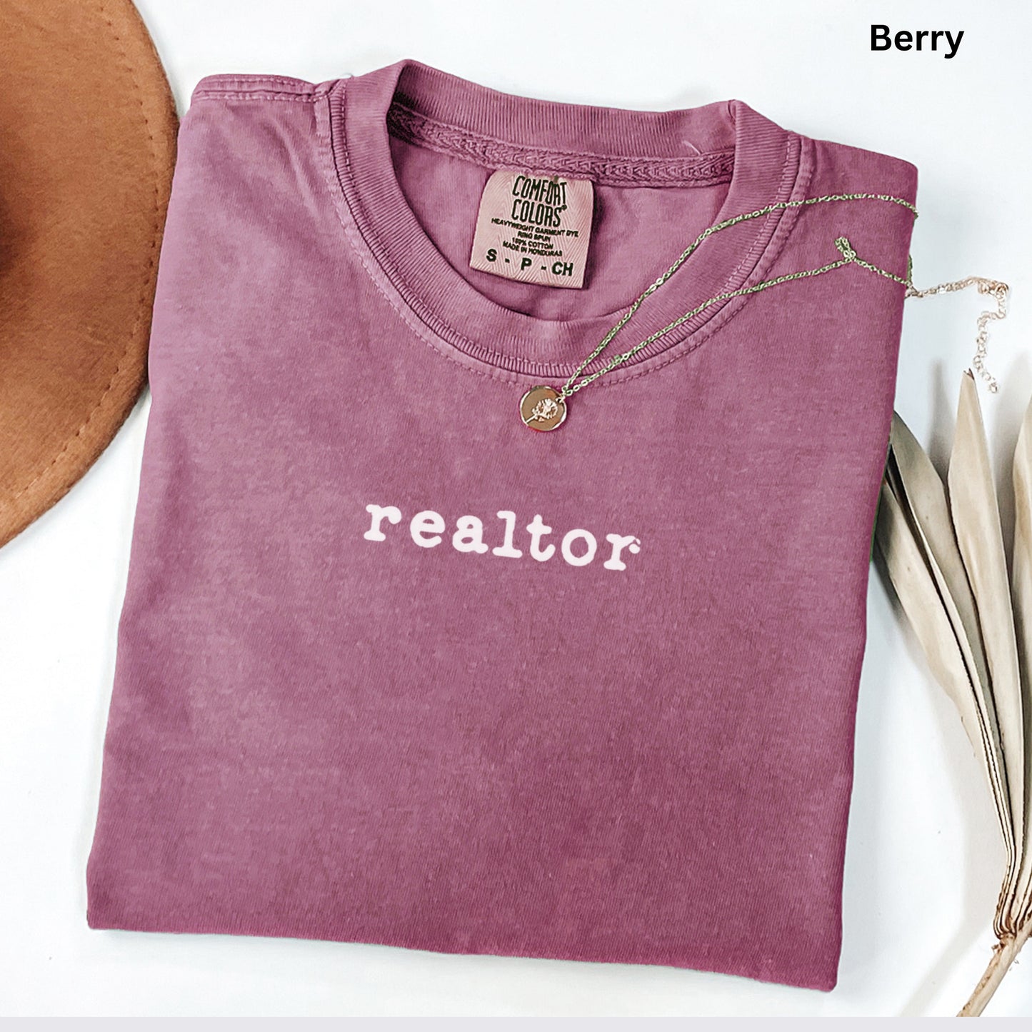 A pink t-shirt with the word "realtor" printed on it, along with a necklace and a brown hat, all laid out on a white surface.
