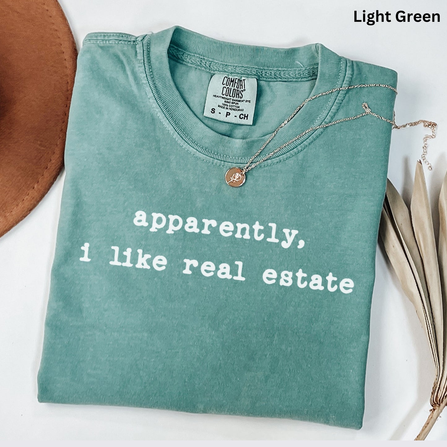 A light green t-shirt with the text "apparently, I like real estate" printed on it, along with a small gold pendant. The t-shirt is laid out on a white surface, and there is a brown hat and some dried leaves nearby.