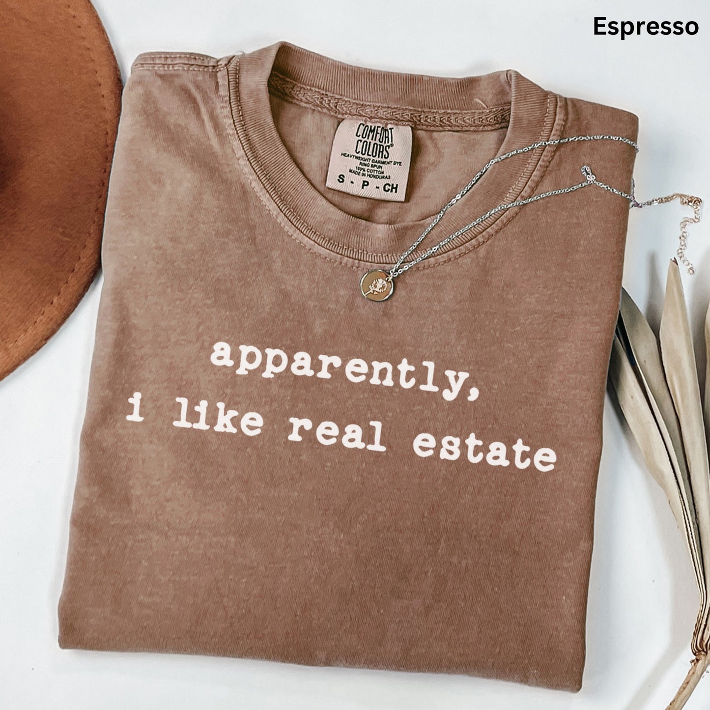 a brown t-shirt with white text that reads "apparently, I like real estate" laid out on a white surface.